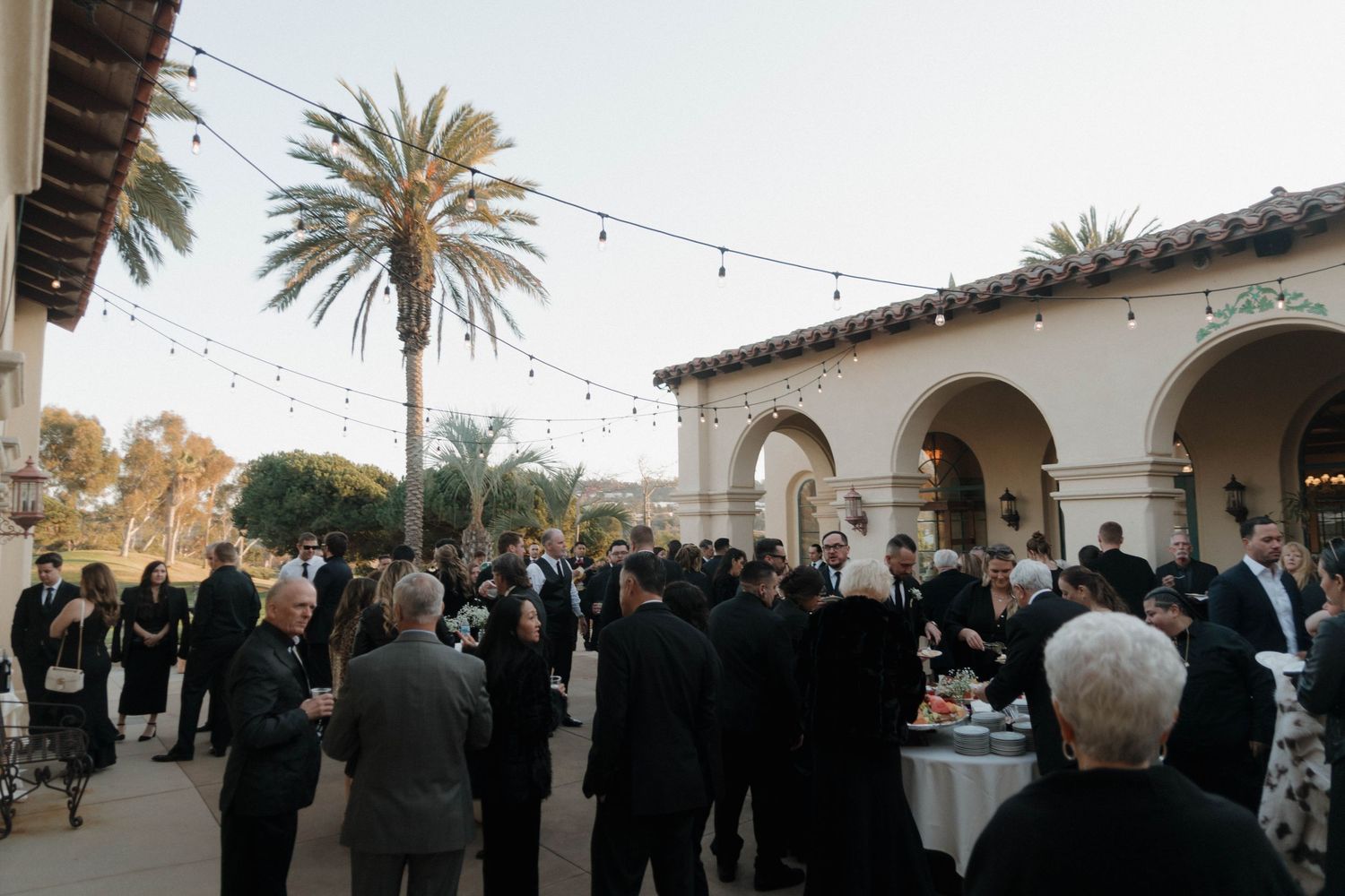 A panoramic view of an outdoor wedding ceremony with string lights and palm trees at sunset.