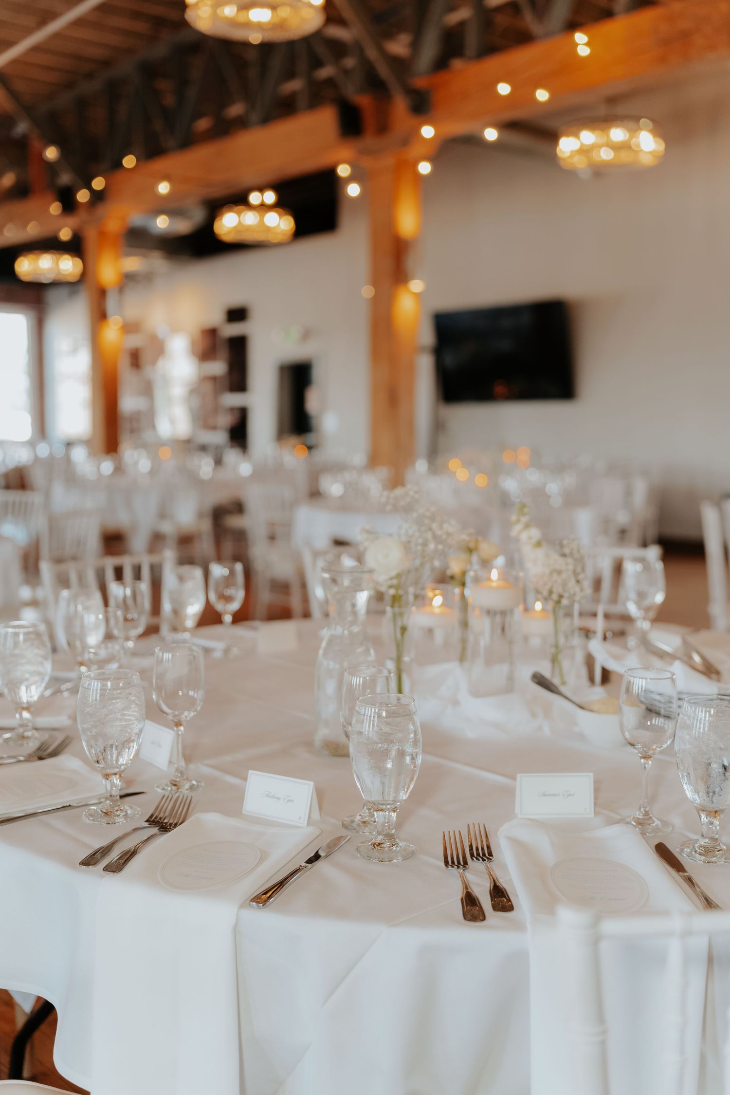 Elegant wedding reception venue with white round tables, floral centerpieces, candles, and warm string lights in a rustic space.