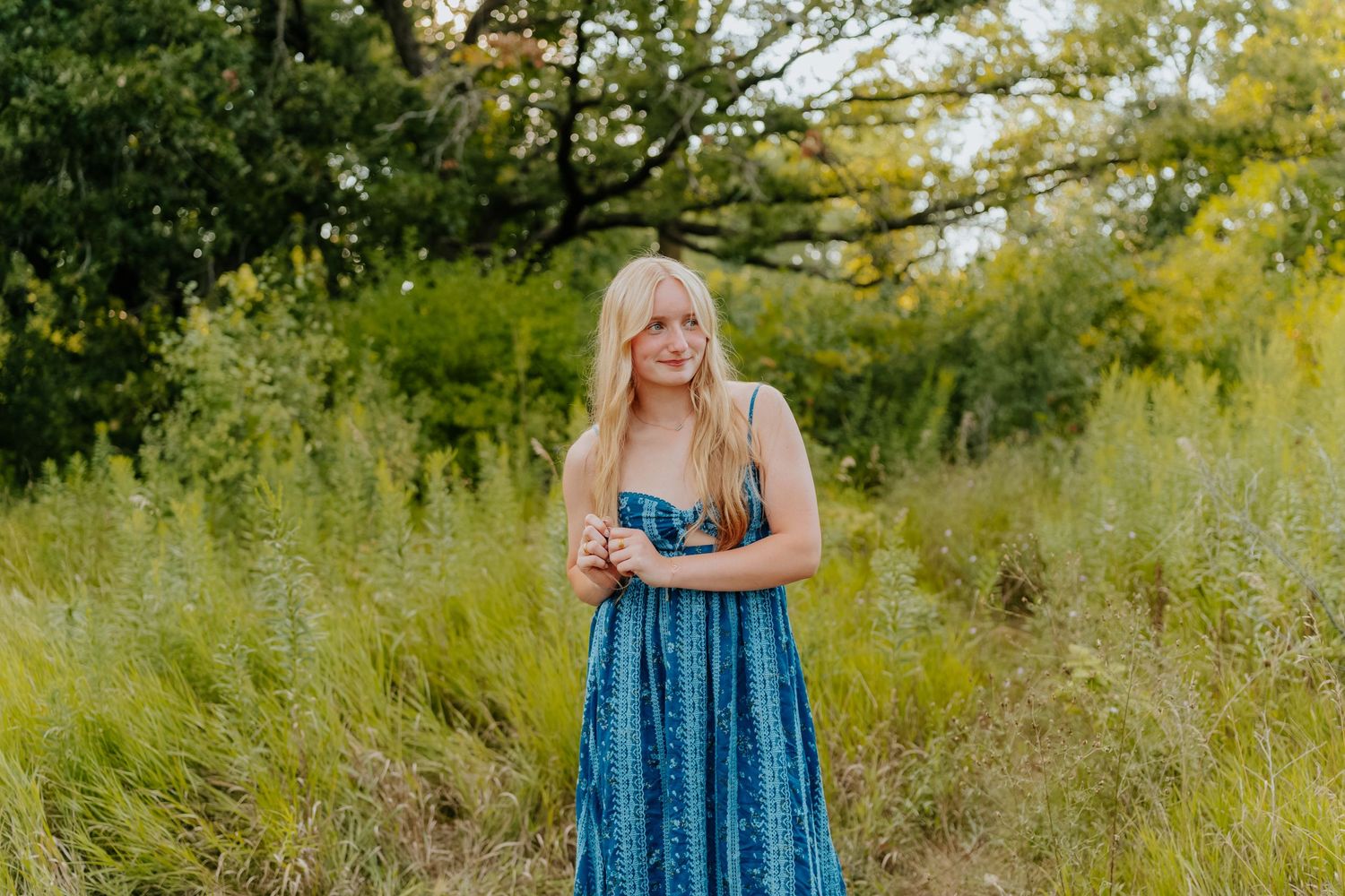 Young blonde woman in a blue maxi dress standing in a lush green meadow with trees in the background.