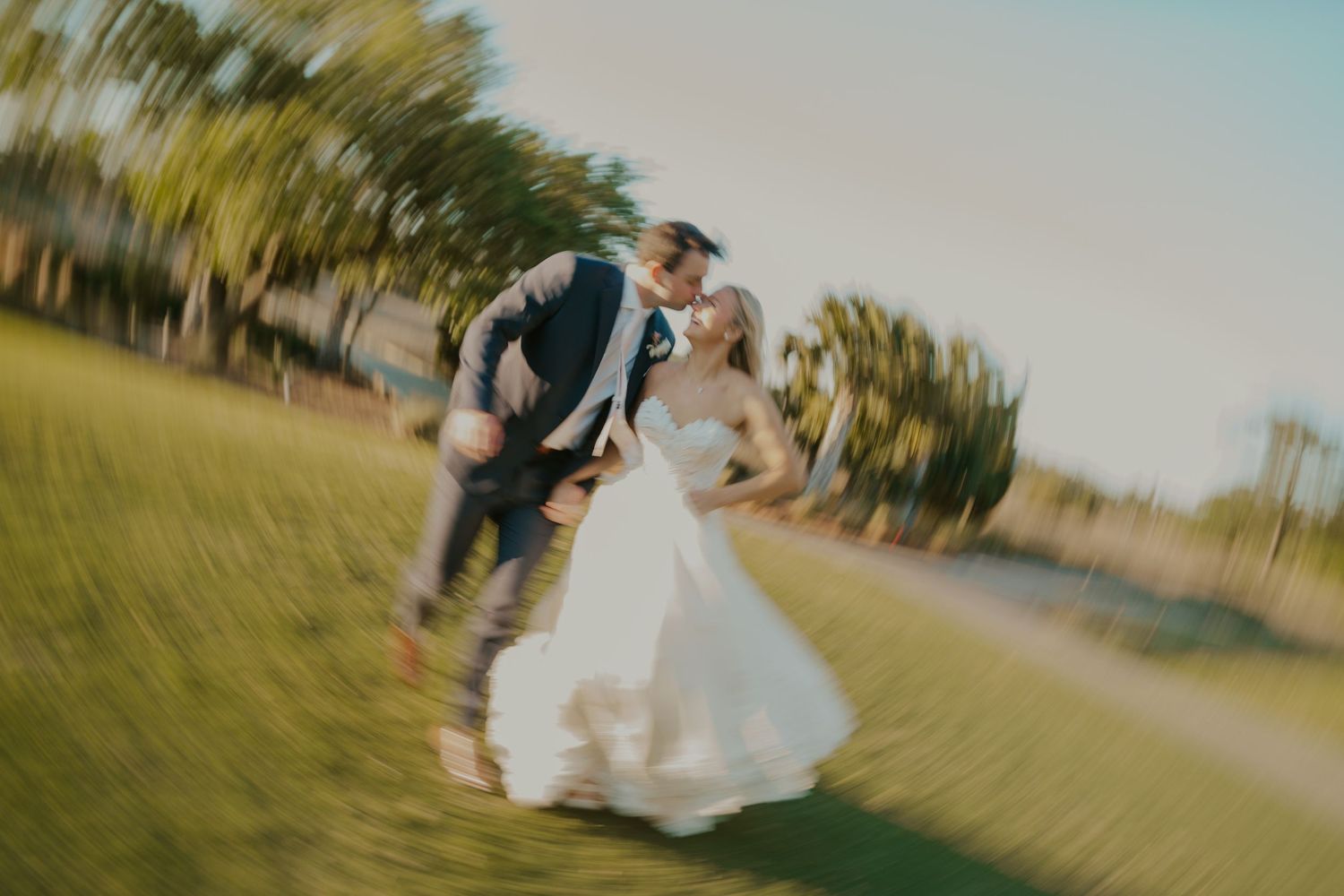 Couple in wedding attire twirls and dances together on a grassy field during sunset.
