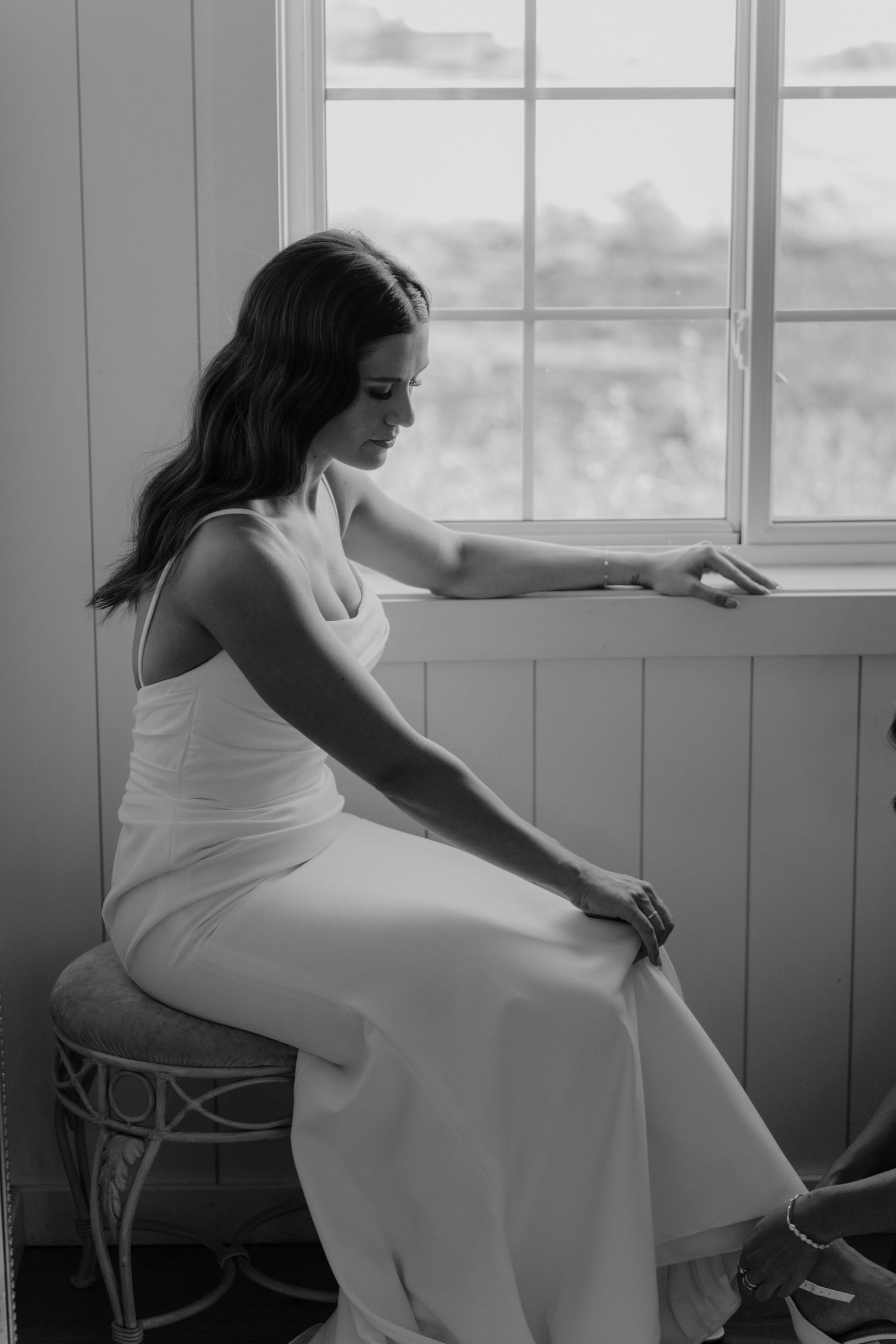 Elegant bride in a white satin gown sitting by a window in a black and white portrait.