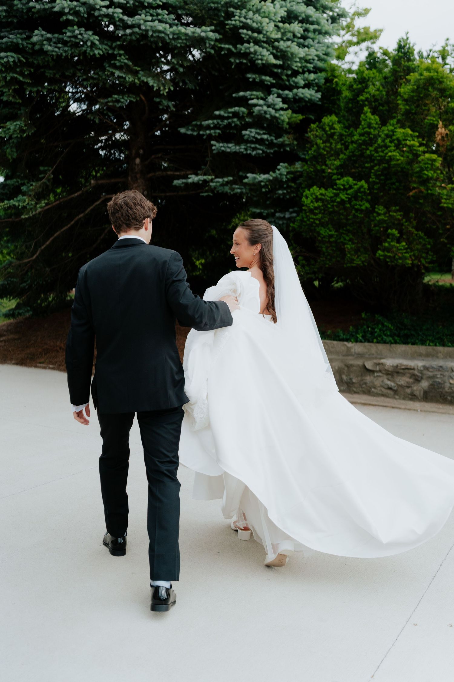 A bride in a flowing white gown and veil walks hand-in-hand with a groom in a black suit outdoors.
