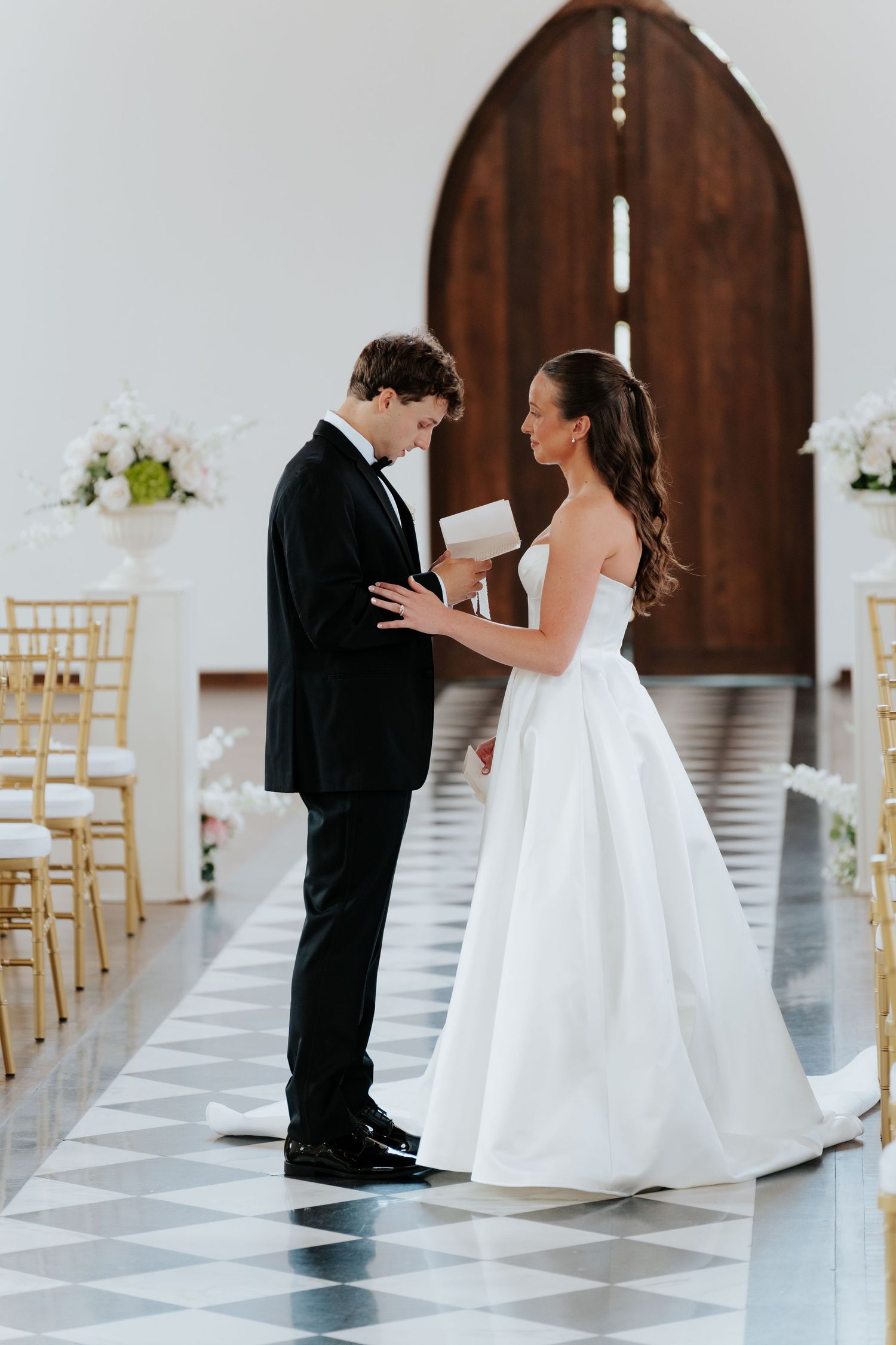 Bride in white gown reads vows to groom in black tuxedo at elegant church wedding ceremony with gold chairs.