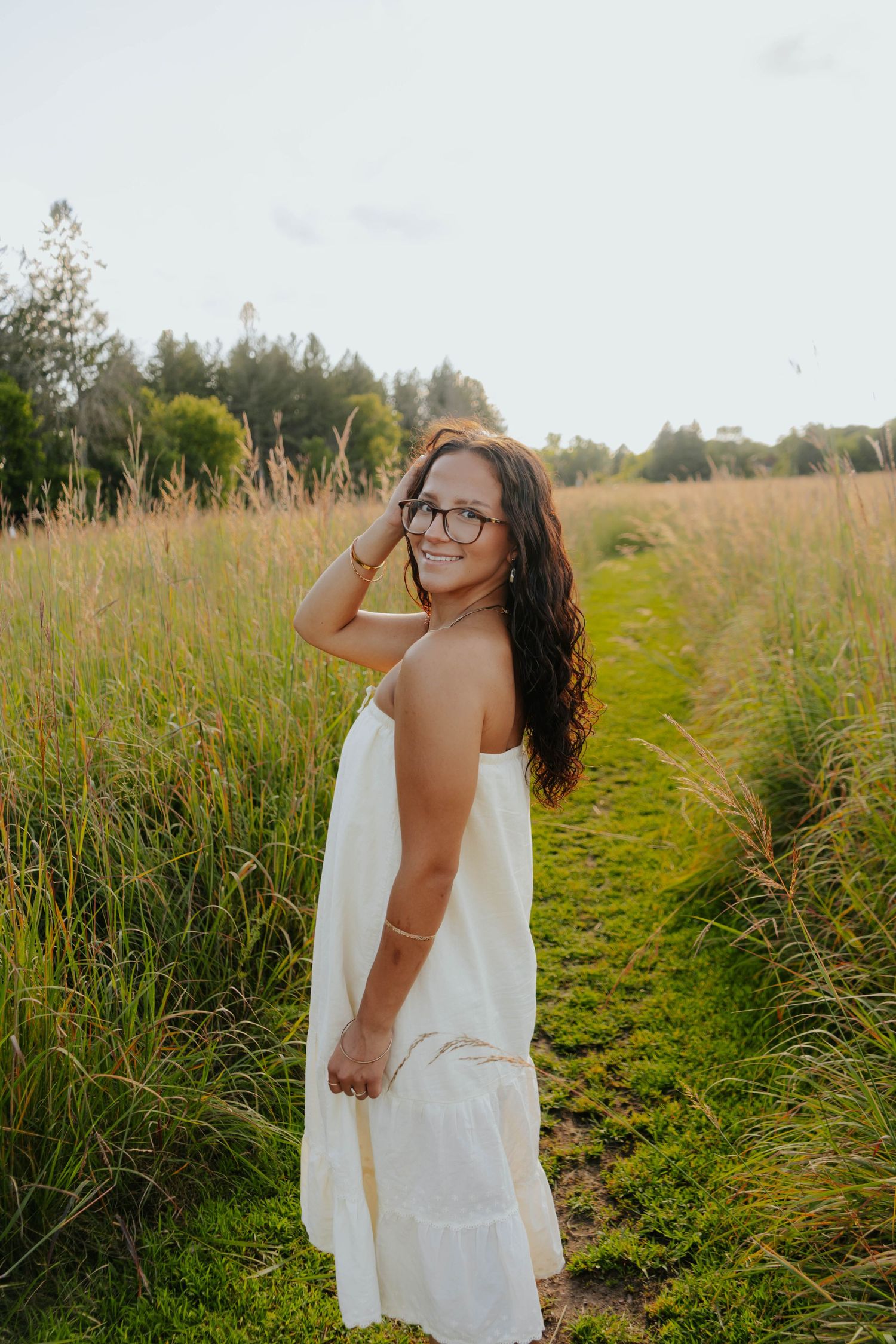 Smiling young woman in white sundress and glasses standing on a grassy path surrounded by tall meadow grass.