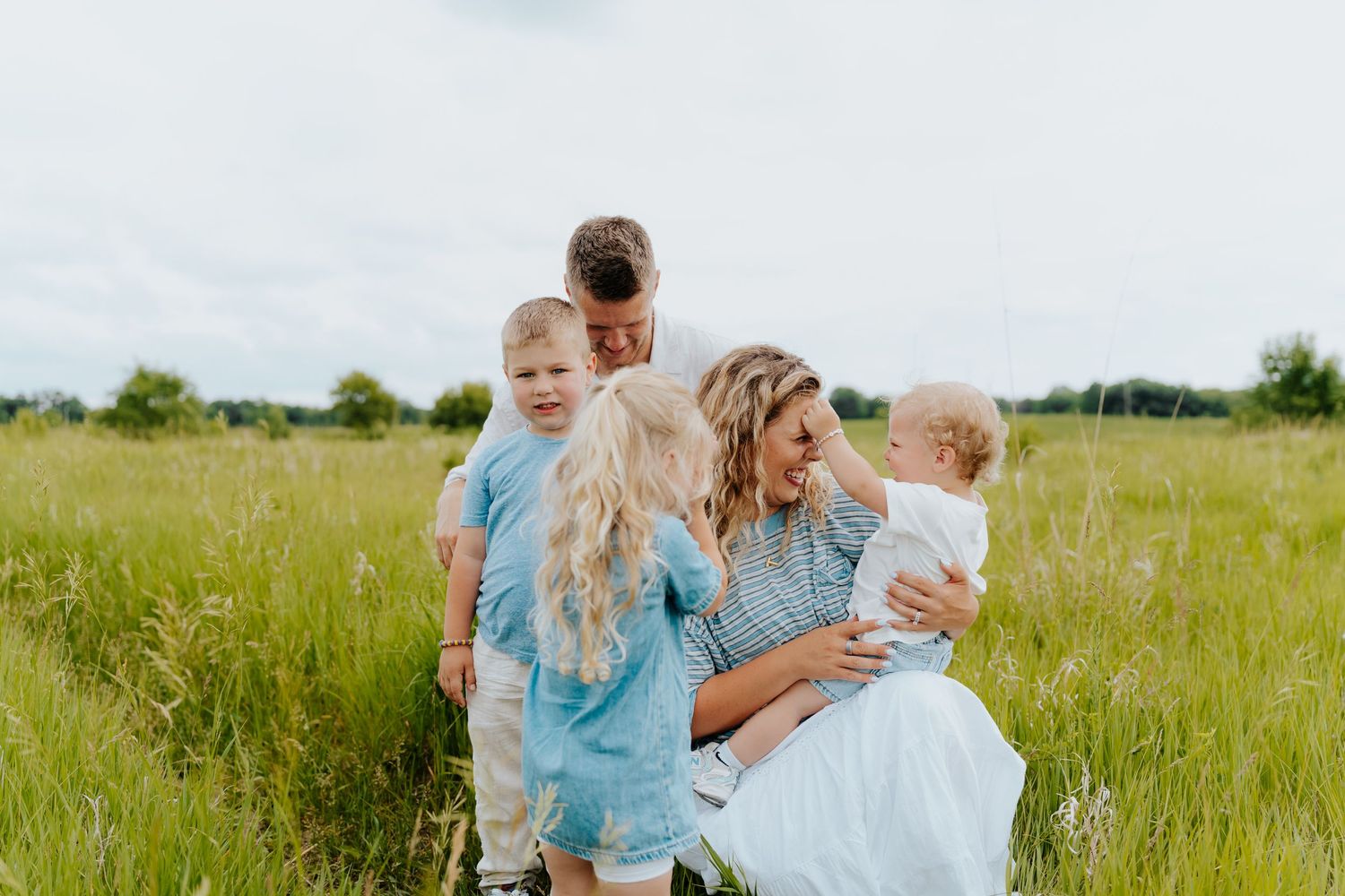 Family of five enjoying a sunny day in a lush green meadow, parents lovingly interacting with three young children.