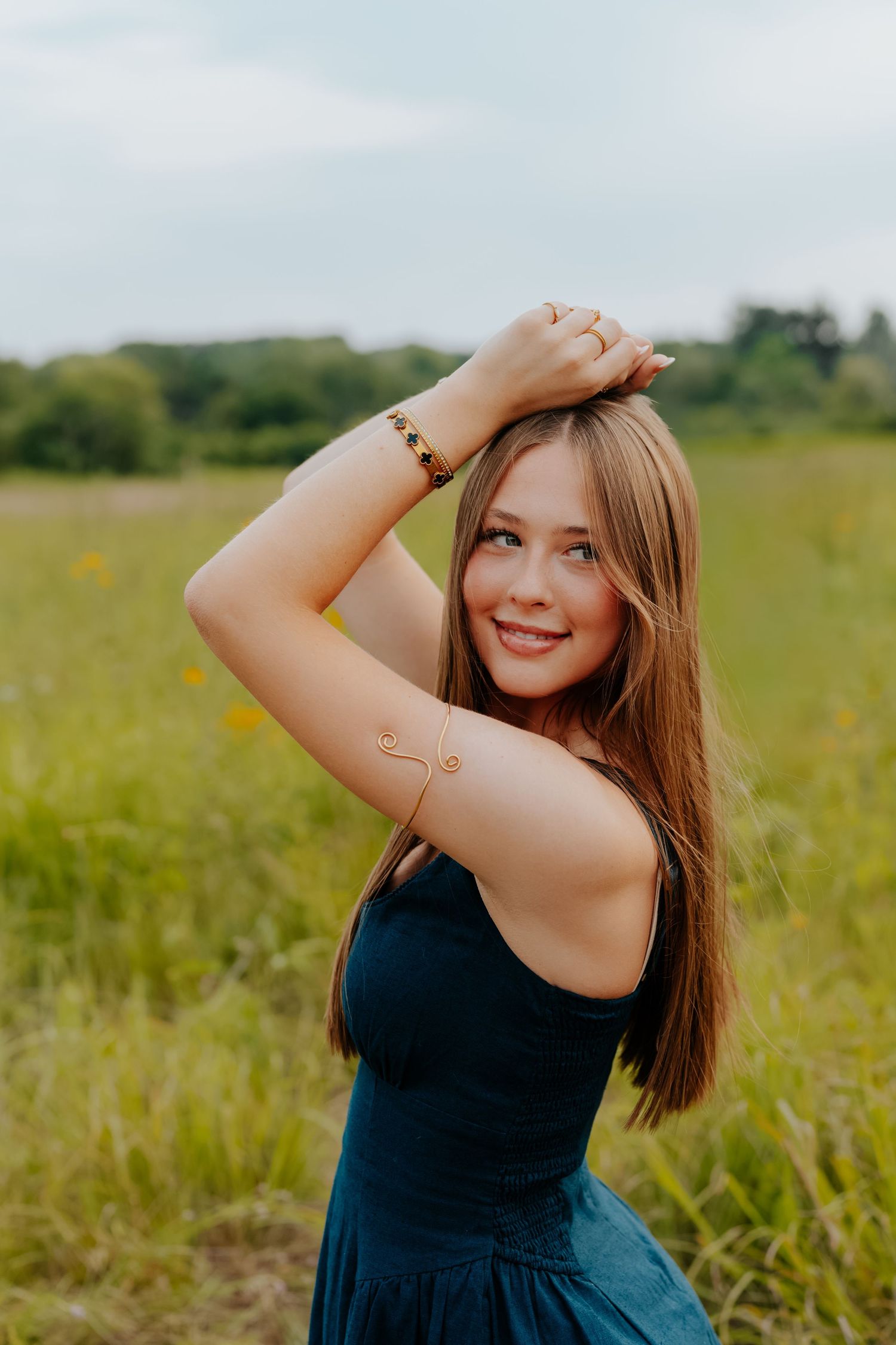 Young woman in teal dress posing in a green field, smiling with hand in hair, wearing gold bracelet.