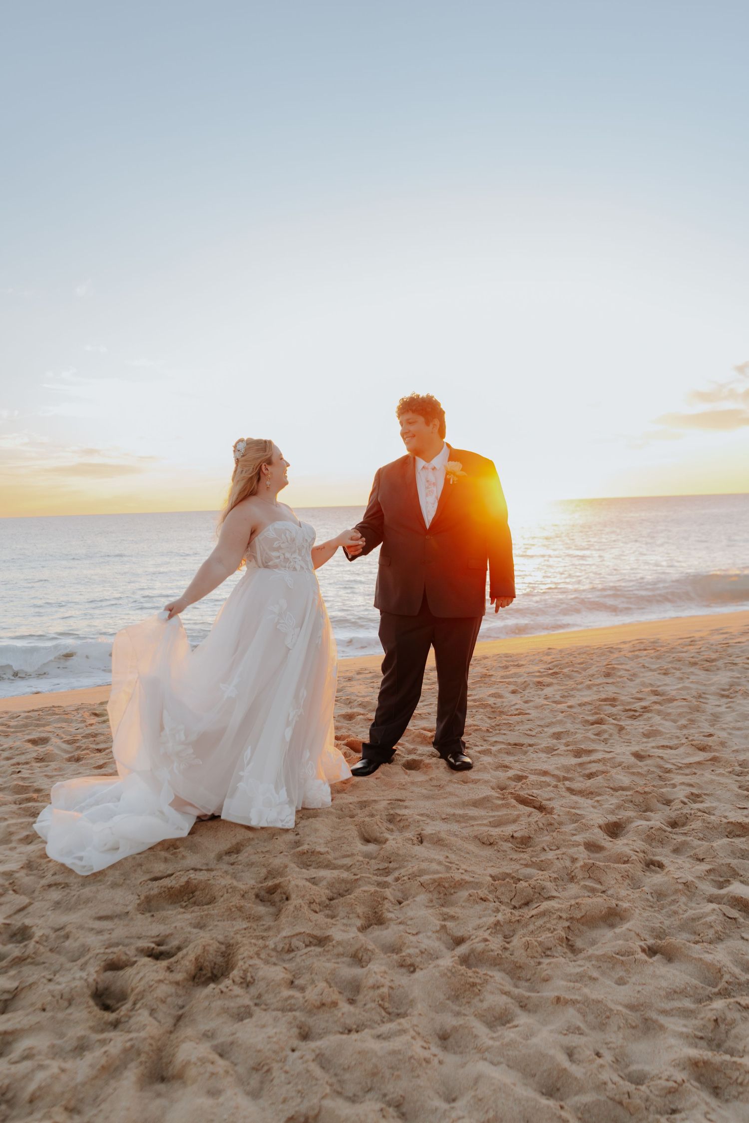 Bride and groom hold hands on sandy beach at golden sunset, bride in flowing white gown, groom in dark suit.