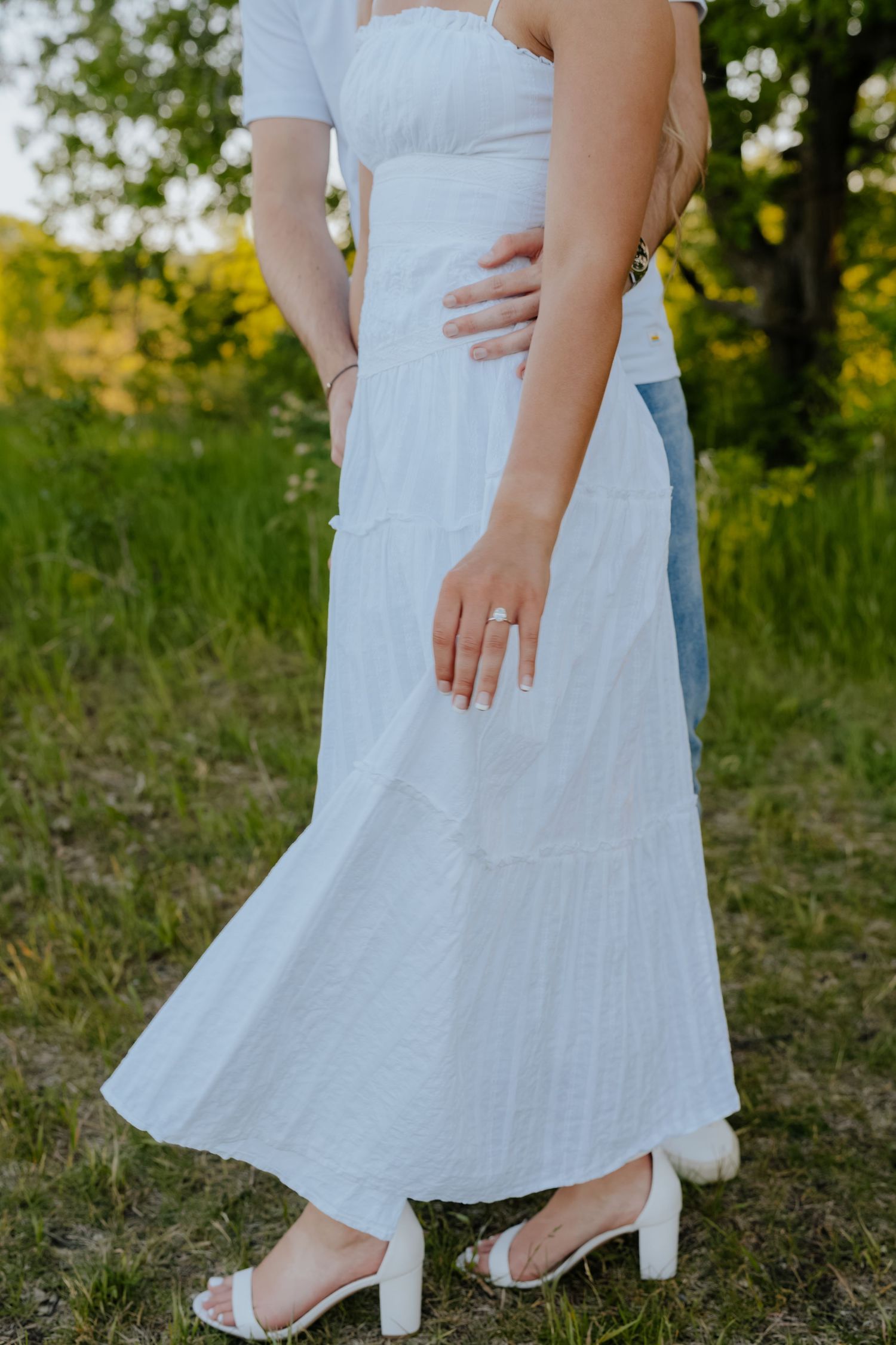 Couple embracing outdoors, woman in white maxi dress and heels with engagement ring, man in jeans, green trees background.