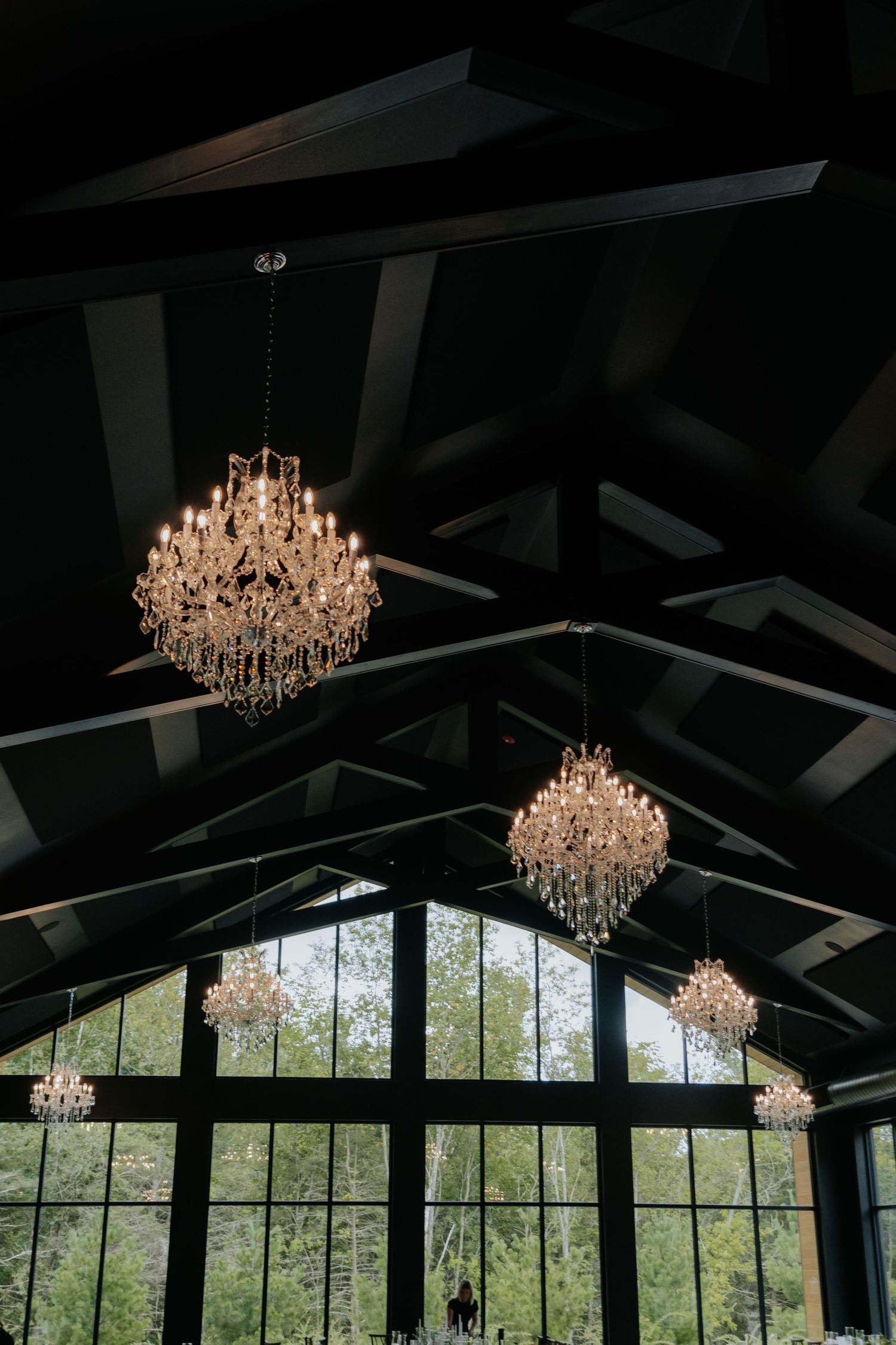 Crystal chandeliers hang from dark vaulted ceiling beams above large black-framed windows overlooking lush greenery.