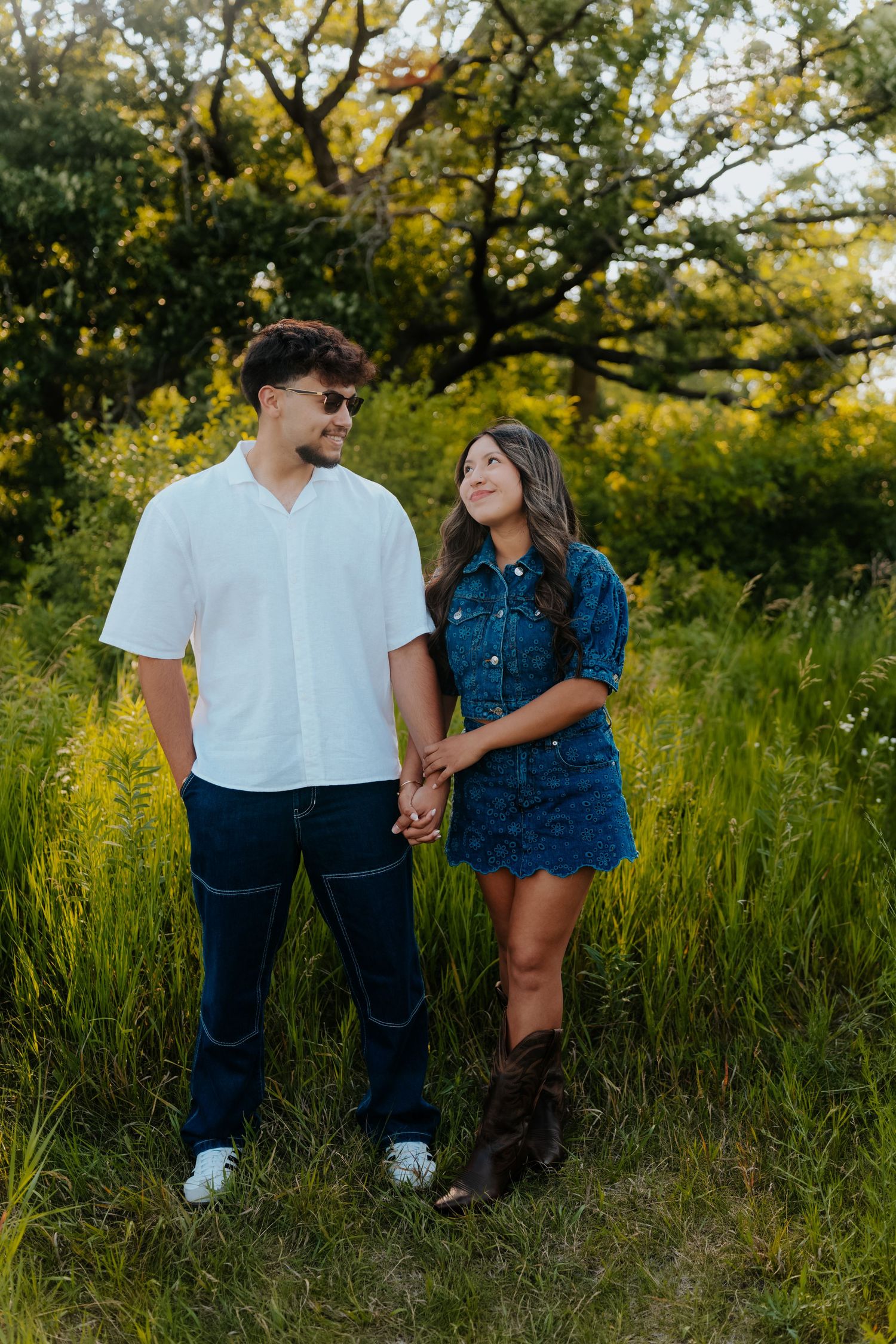 Couple holding hands in a lush green field, man in white shirt and jeans, woman in denim dress and cowboy boots.