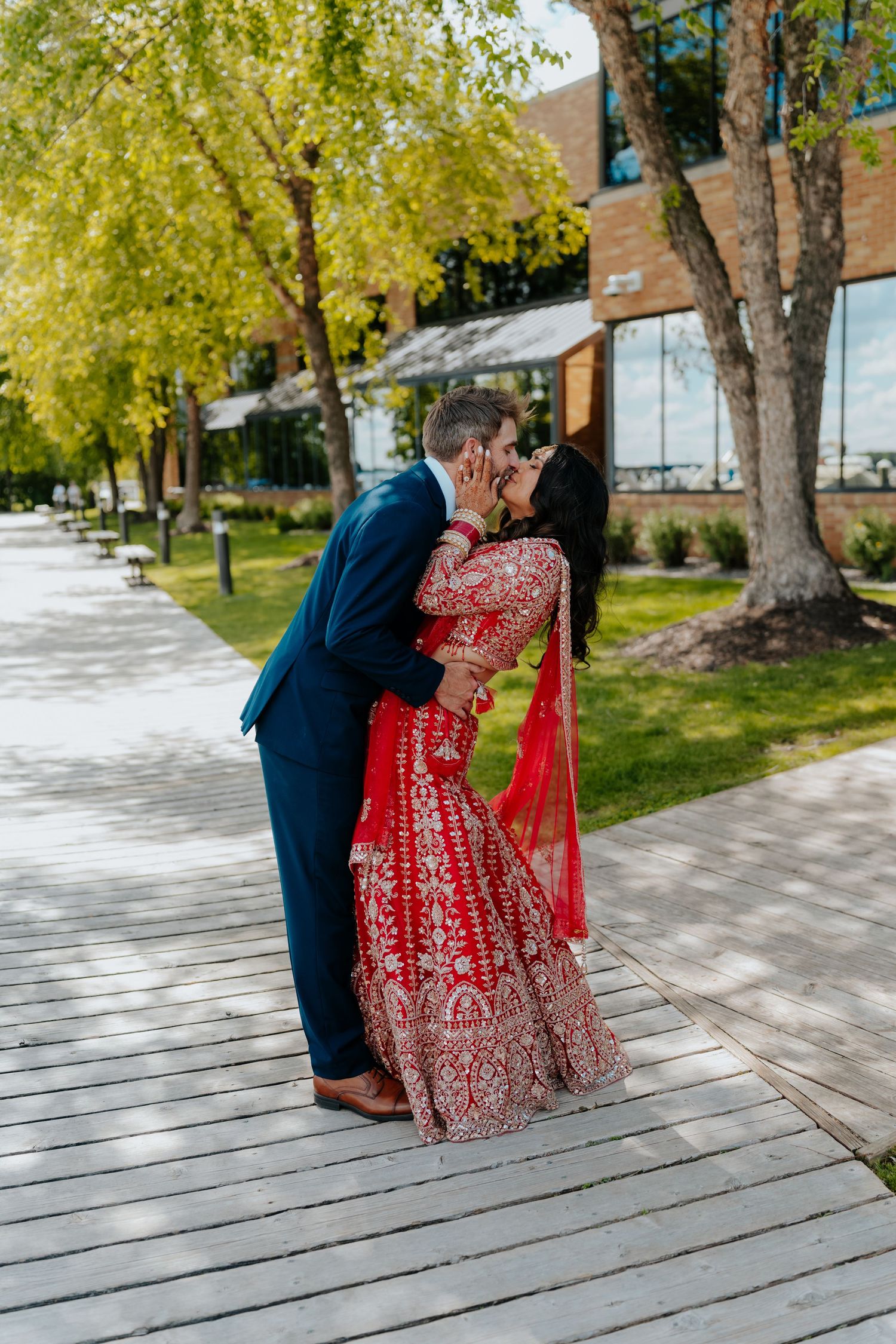 Couple sharing a romantic kiss outdoors, bride in red Indian bridal lehenga and groom in navy suit on wooden path.
