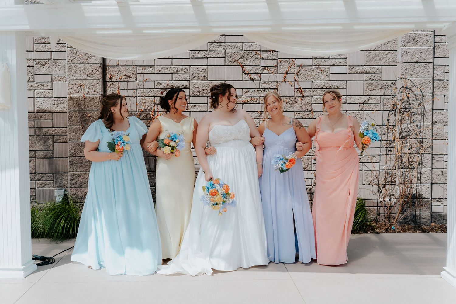Bride in white gown poses with four bridesmaids in pastel blue and pink dresses holding bouquets at outdoor wedding venue.
