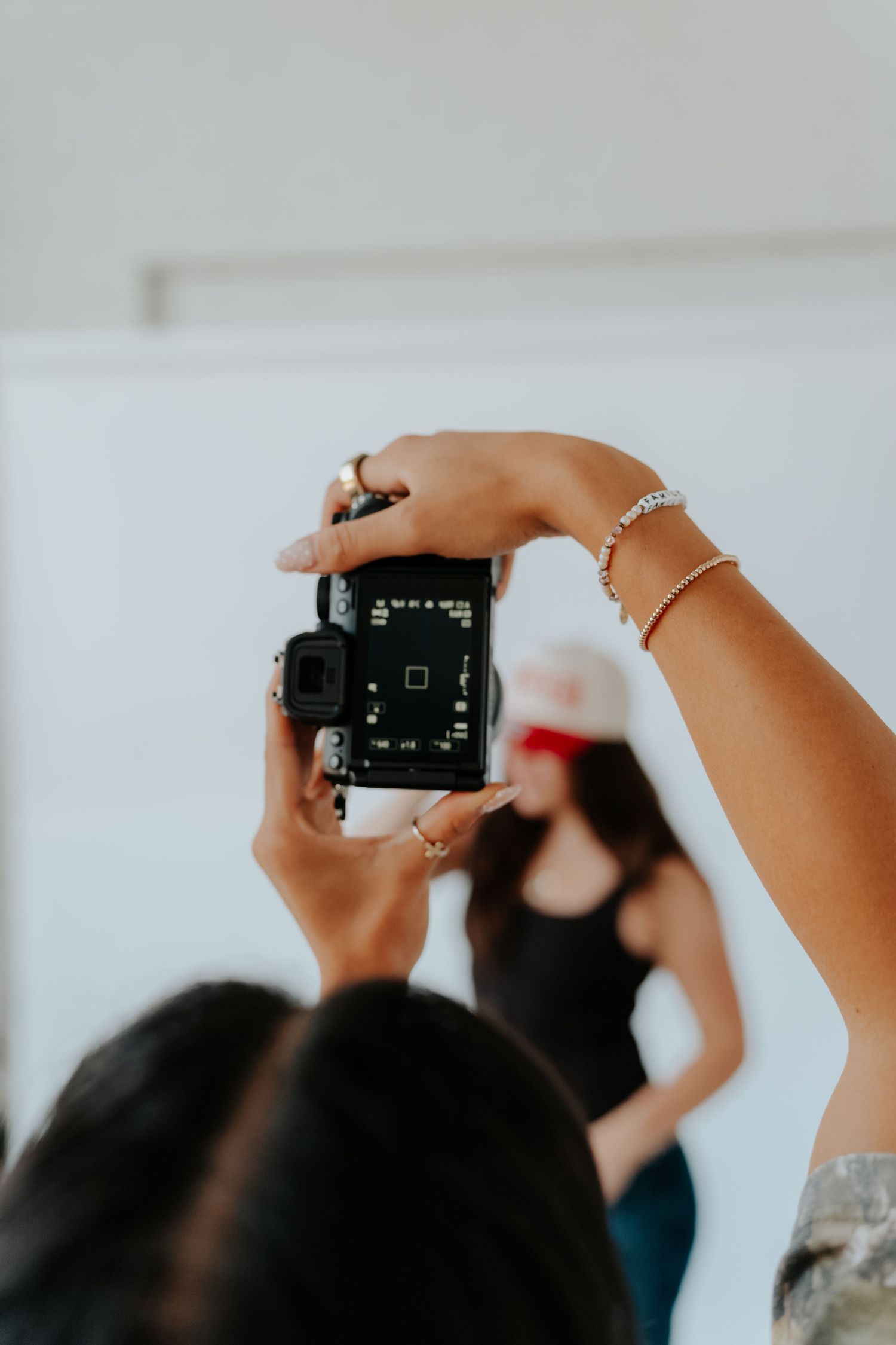 Photographer holds DSLR camera overhead to capture a woman in black top and red cap in a bright studio.