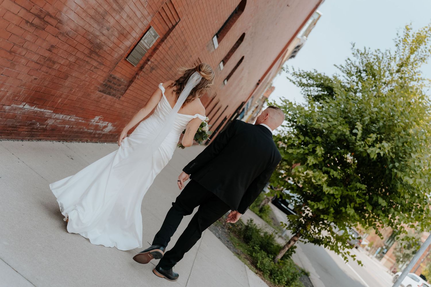 Bride in white gown and groom in black suit walk hand in hand past a brick building on their wedding day.