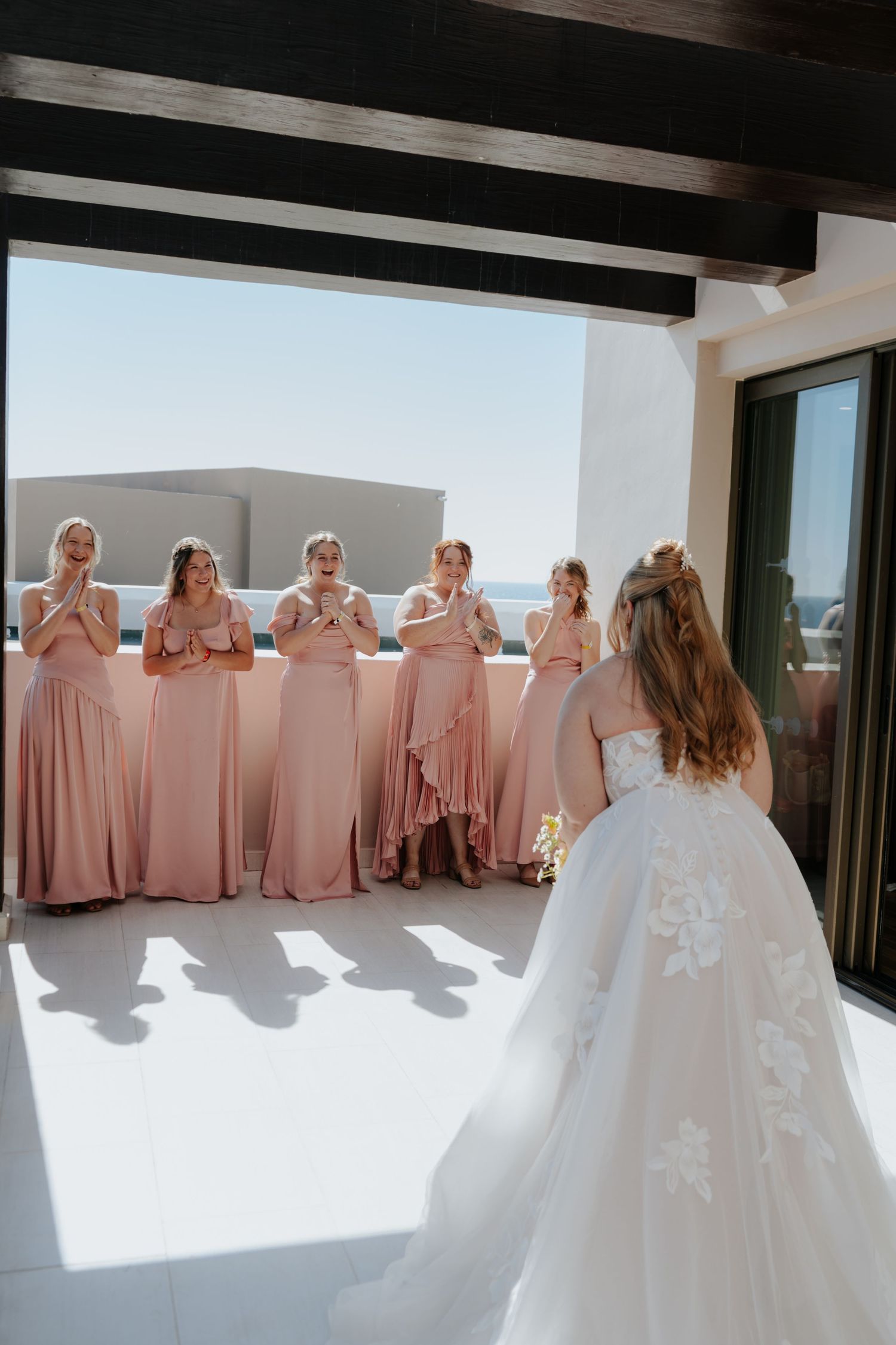 Bride in white lace gown reveals herself to bridesmaids in pink dresses who react with joy on a sunlit terrace.