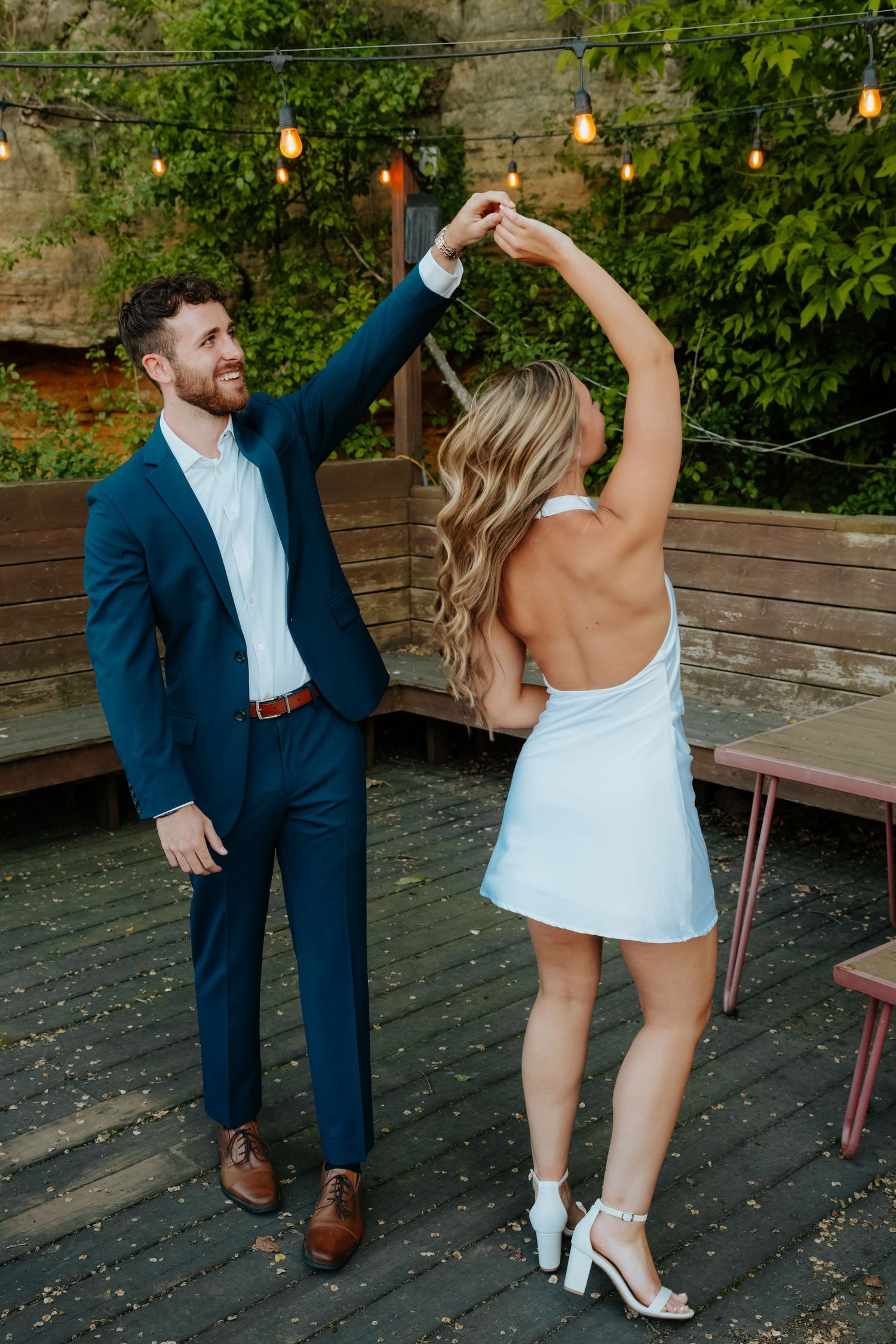 A groom in a navy suit twirls his bride in a white backless mini dress on an outdoor patio with string lights.