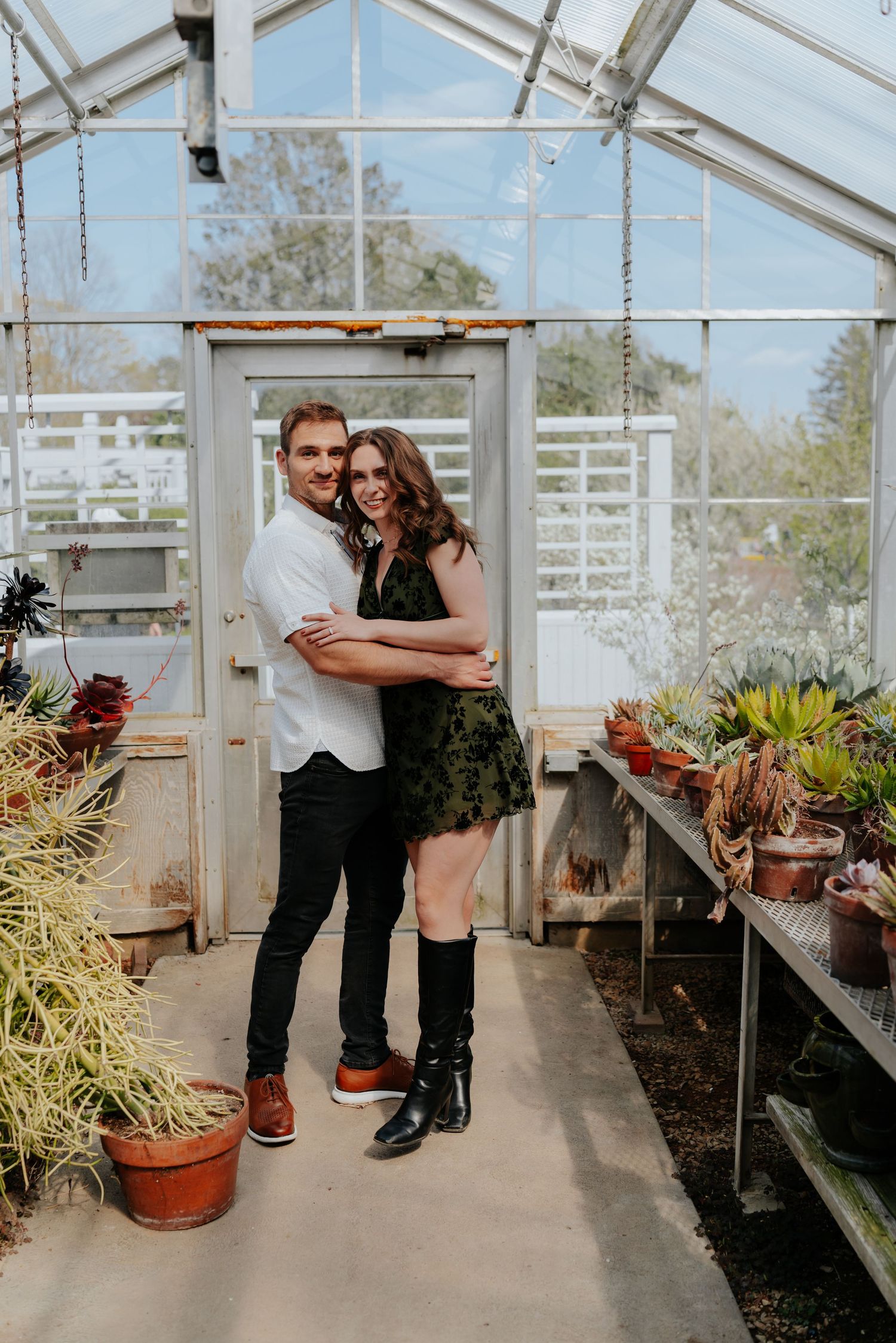 Couple embracing and smiling in a sunlit greenhouse surrounded by potted succulents and plants.