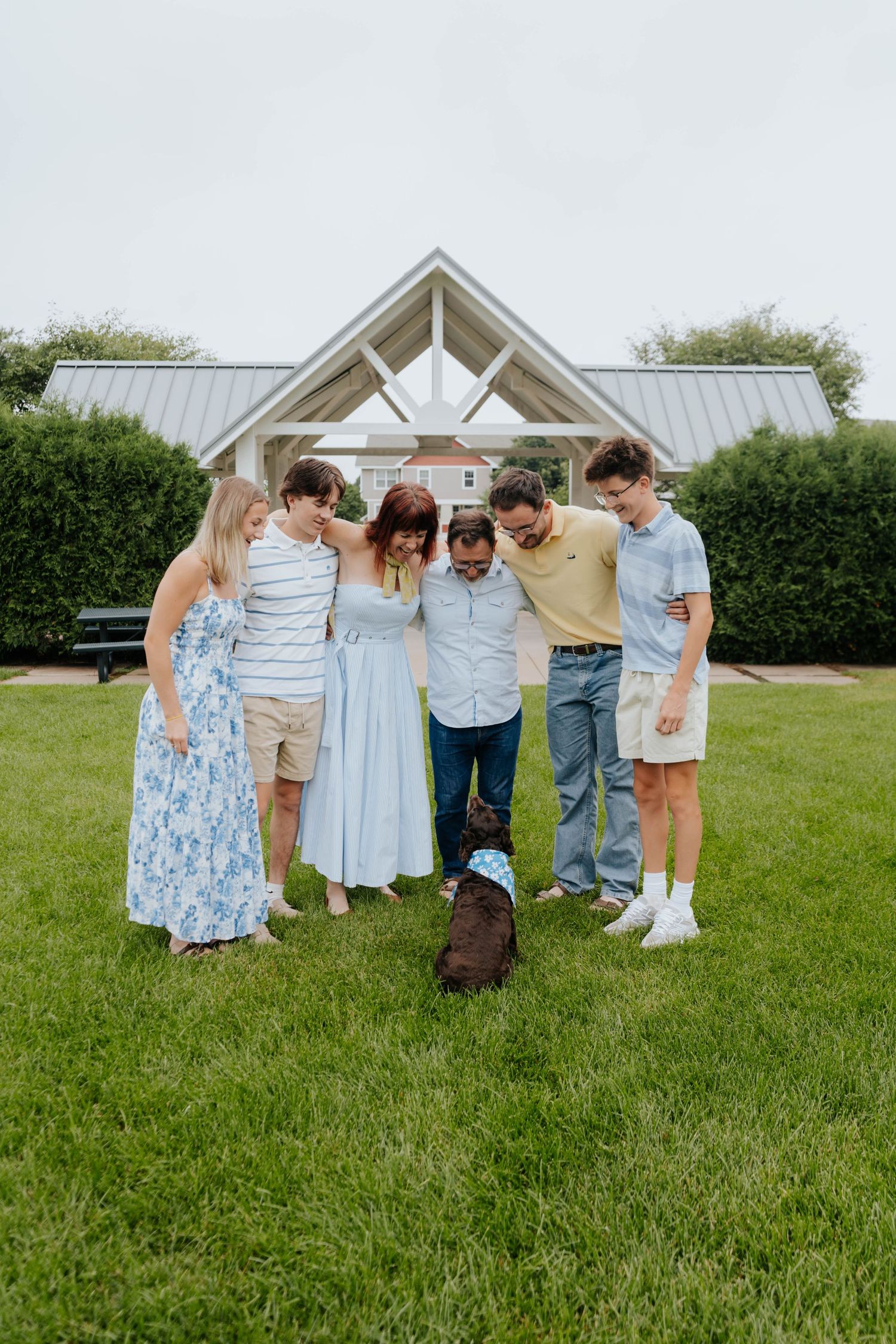 Family of five huddles together on green lawn with their dog near a white pavilion outdoors.