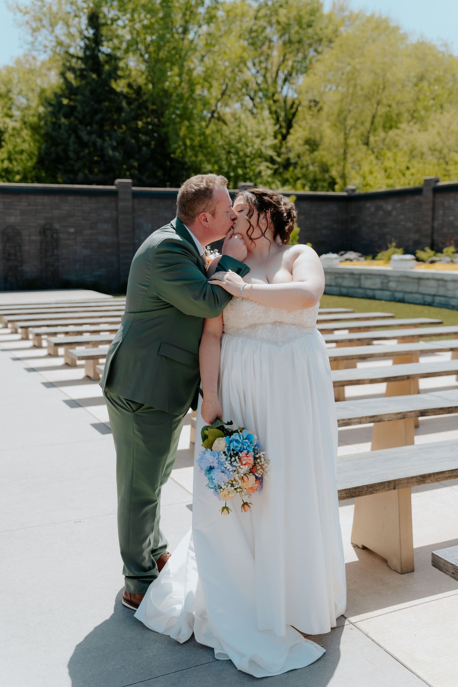 Bride and groom share first kiss in outdoor wedding ceremony, bride holding colorful bouquet, groom in green suit.