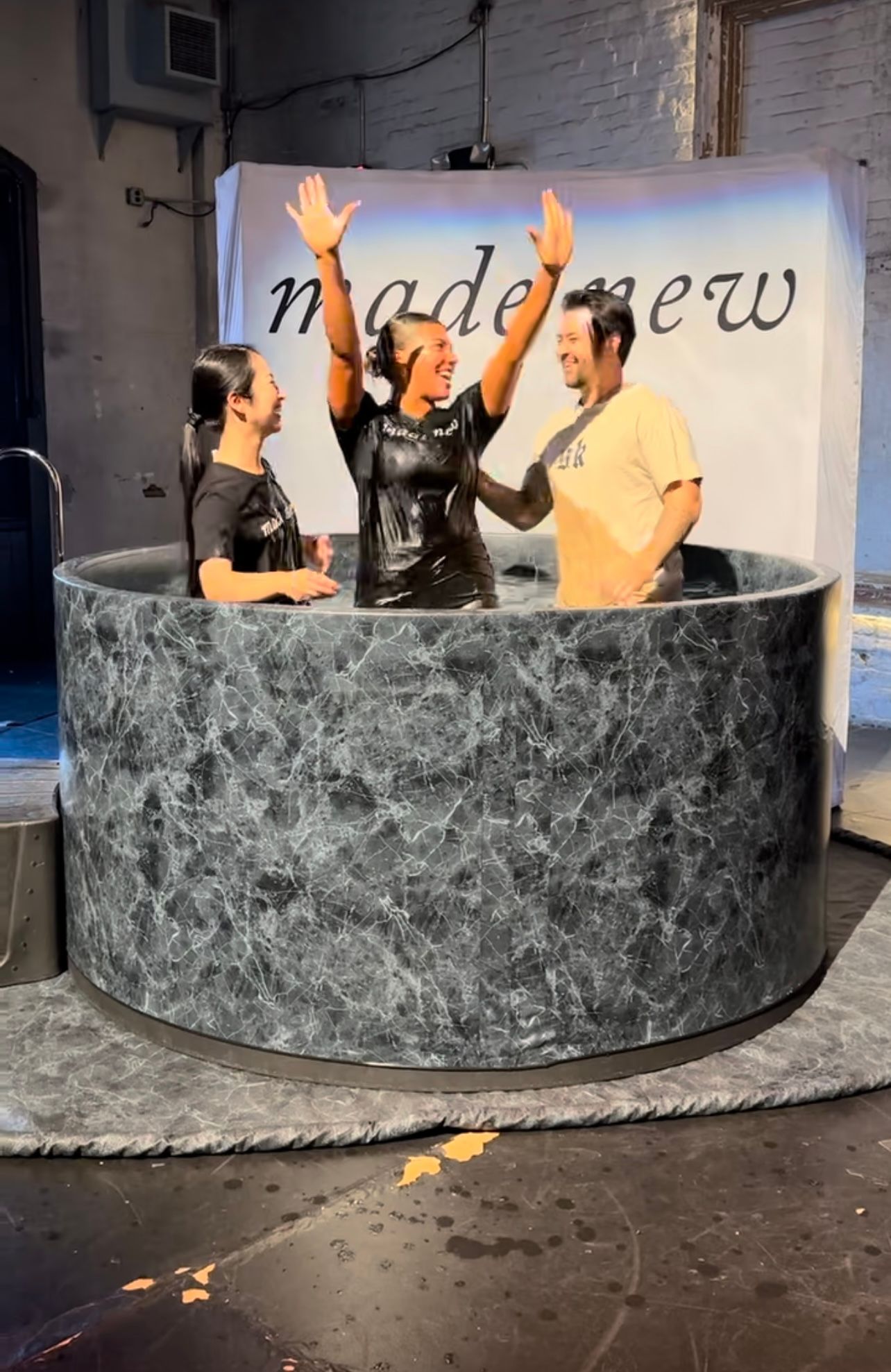 People raise their hands joyfully while sitting in a circular marble-textured structure in a studio space.