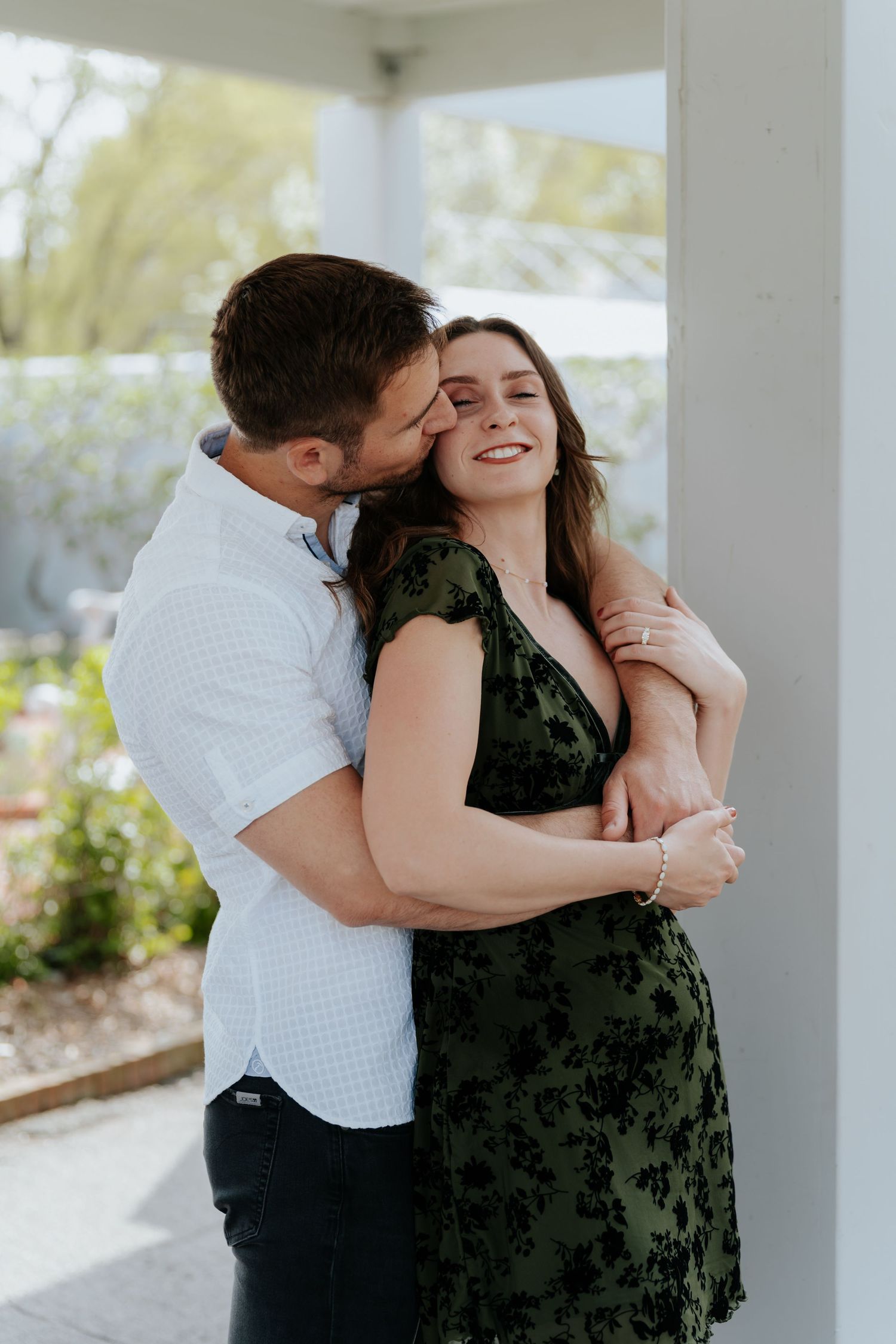 A couple shares a tender embrace on a porch, man kissing woman's cheek as she smiles in a green floral dress.