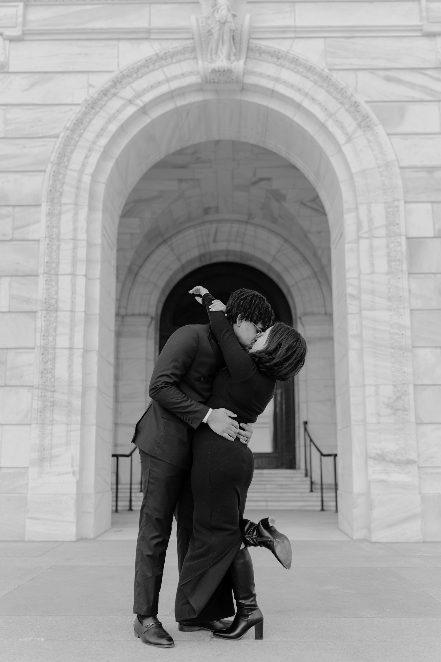 Black and white photo of a couple kissing passionately in front of a grand stone archway entrance.