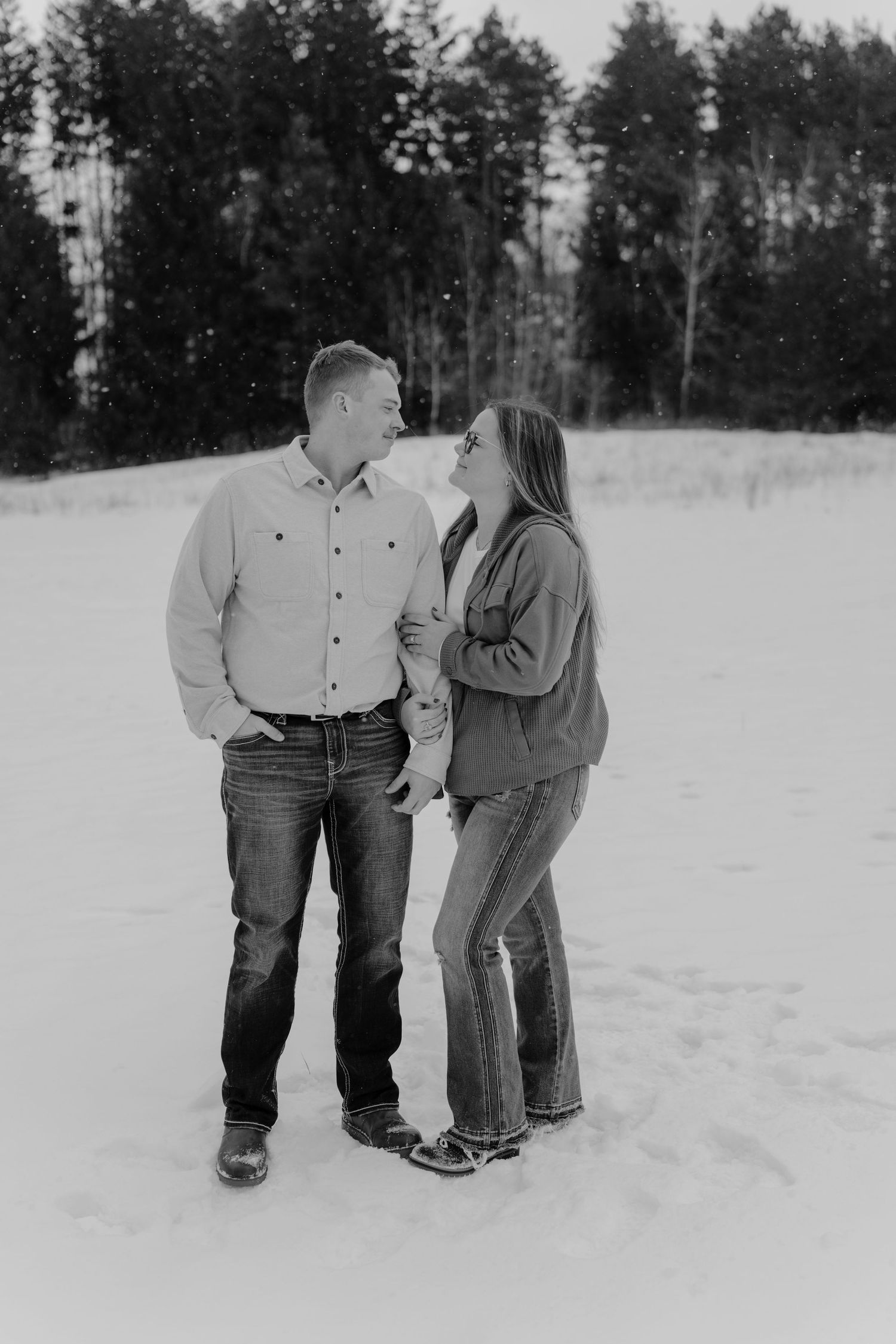 Black and white photo of a couple standing in snow, smiling at each other with evergreen trees in background.
