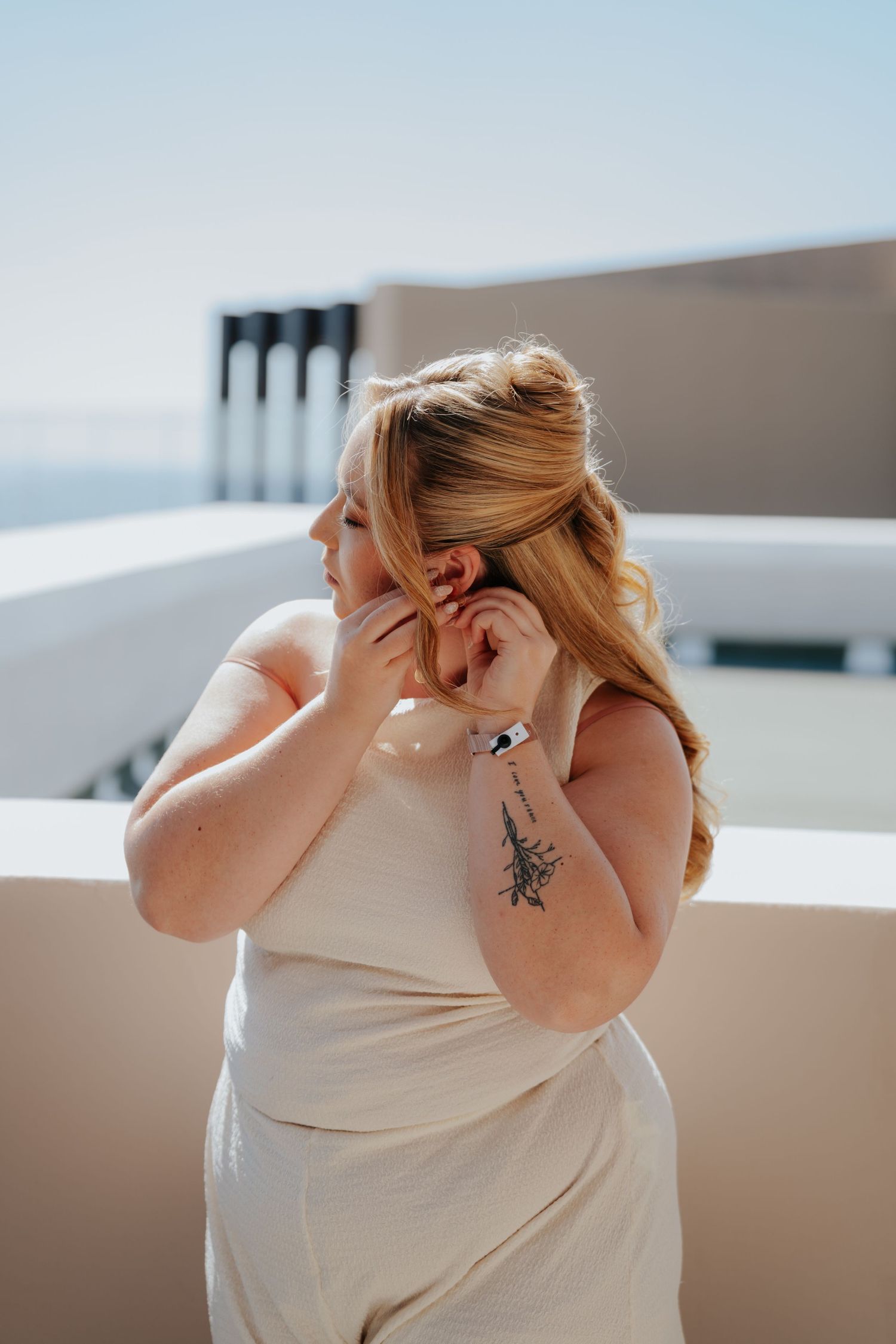 Blonde woman in white dress adjusting earring on sunny balcony, tattoo visible on hand.
