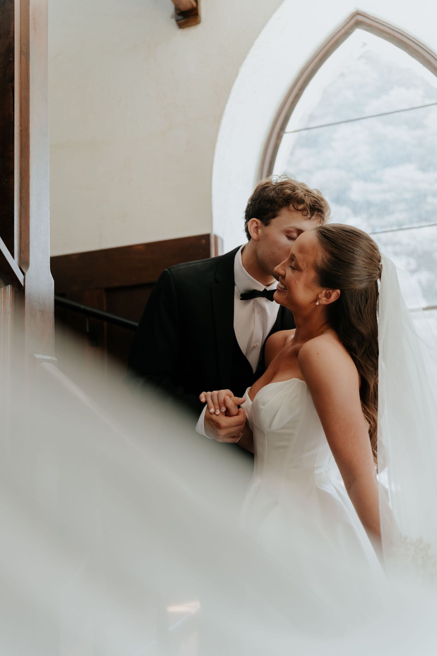 Bride and groom share a romantic kiss in a sunlit church, dressed in a white gown and black tuxedo.