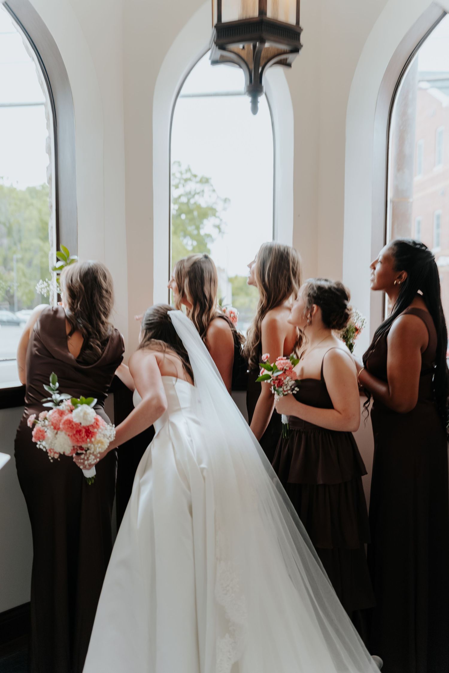 Bride in white gown with bridesmaids in dark dresses holding pink bouquets, standing by arched windows in a chapel.
