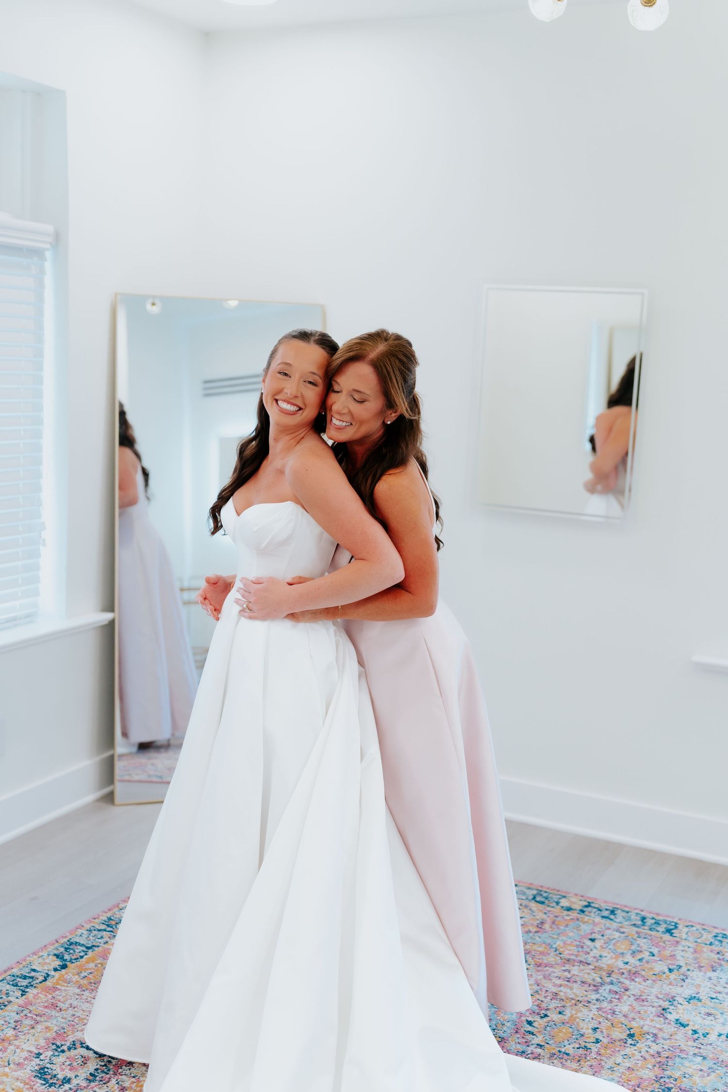 Bride in white strapless gown hugging bridesmaid in blush pink dress, smiling in bright dressing room with mirrors.