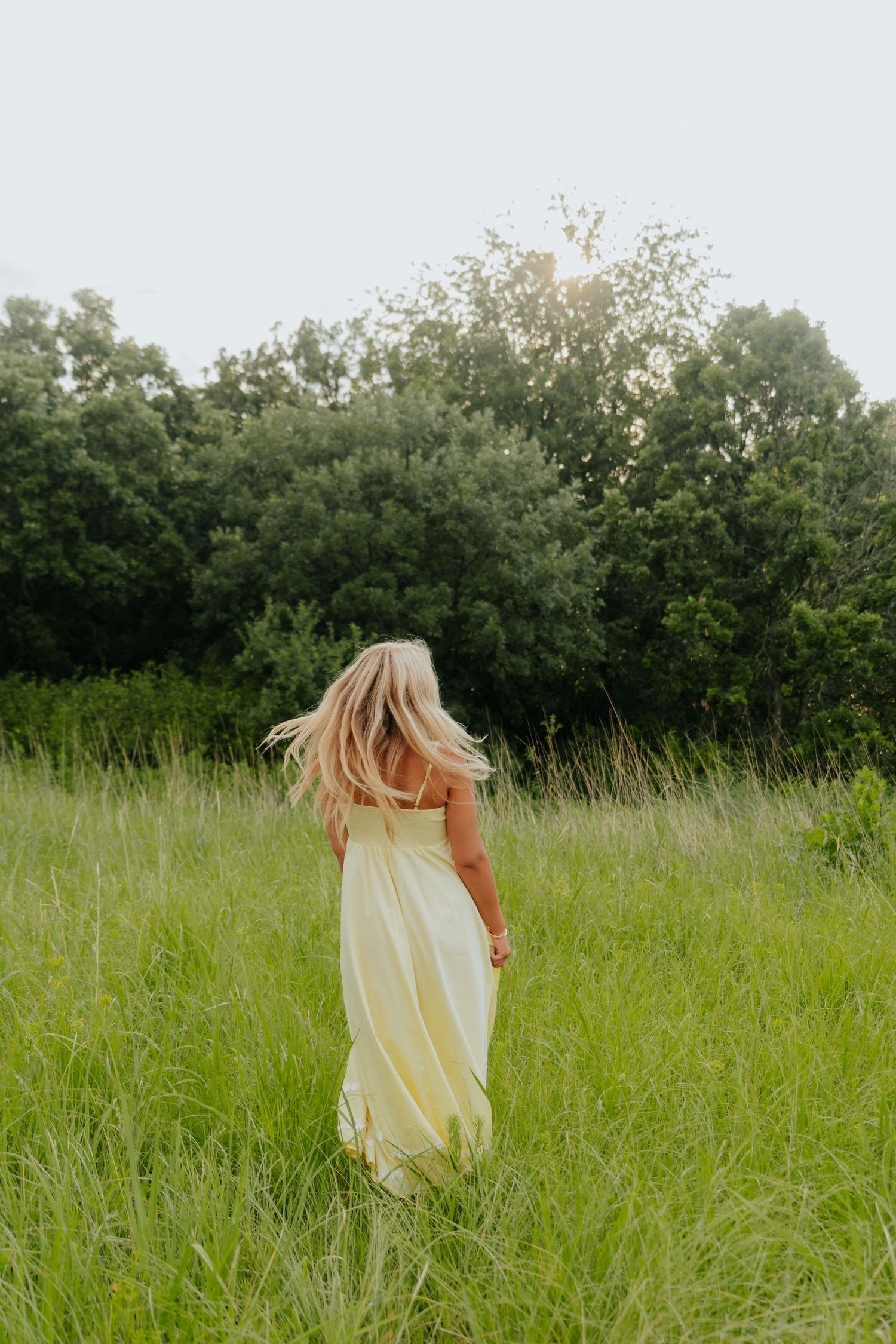 Blonde woman in a flowing yellow maxi dress walking through a lush green meadow with trees in the background.