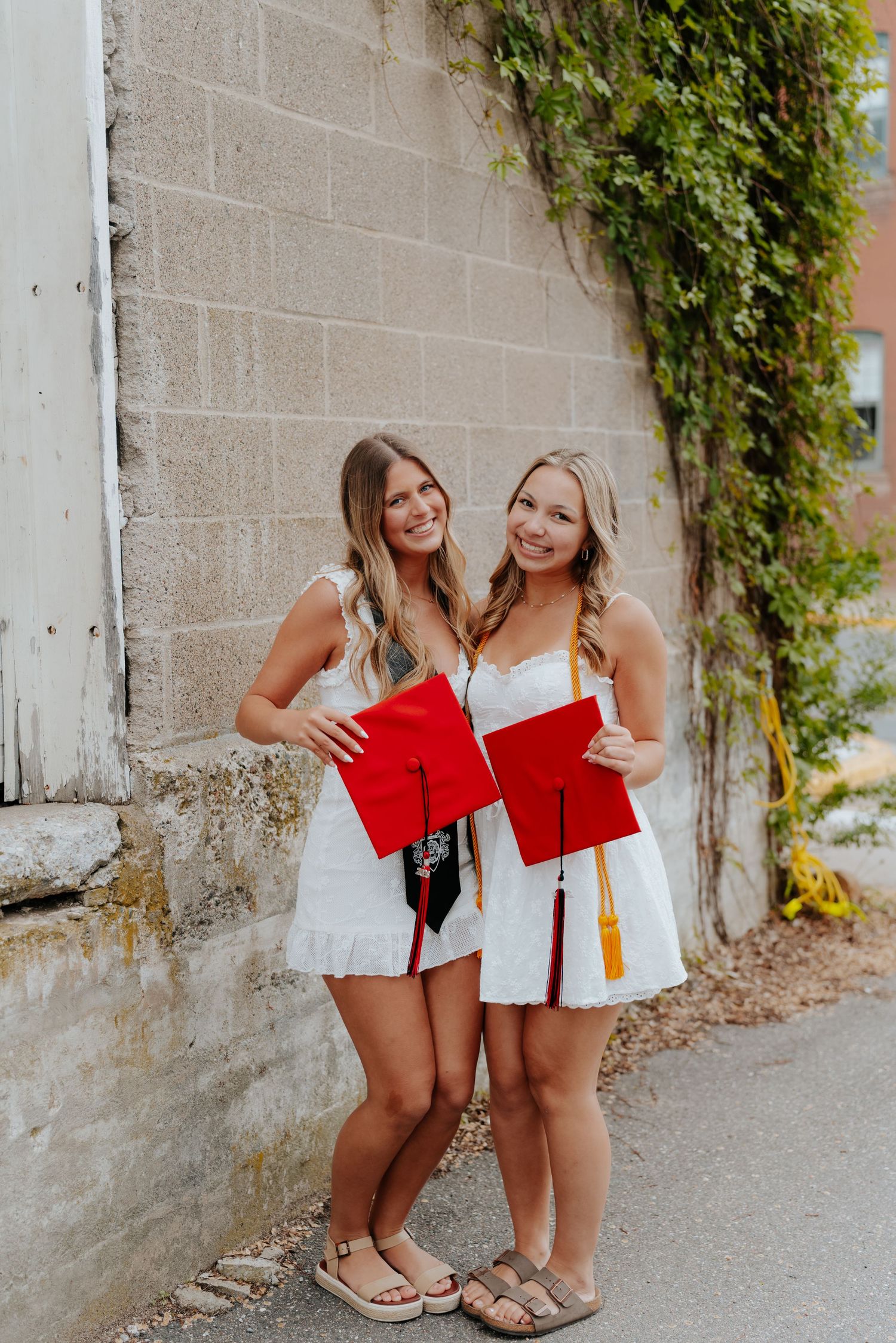 Two smiling female graduates in white dresses holding red graduation caps against a brick wall.