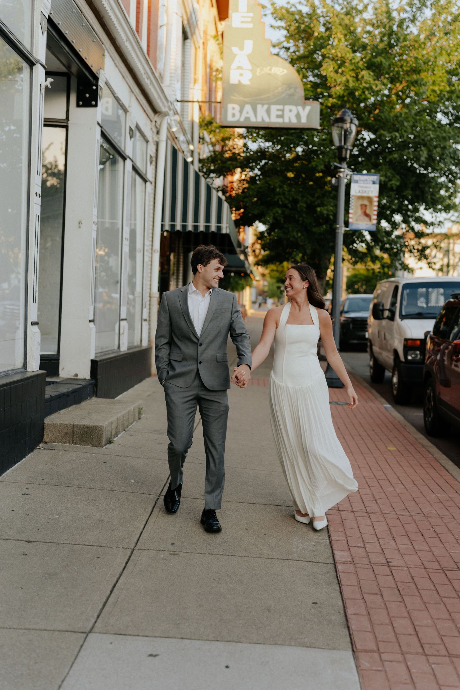 A bride and groom walk hand in hand on a brick sidewalk past a bakery storefront on a sunny day.
