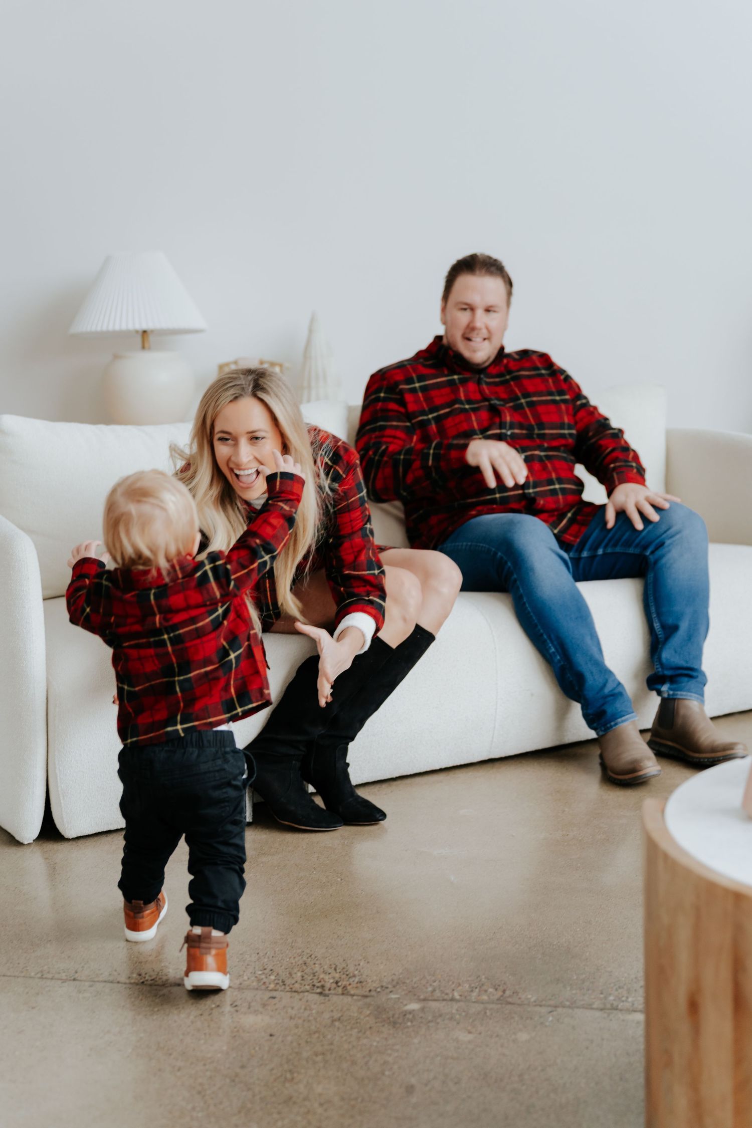Family of three in matching red plaid shirts sitting on white couch, toddler running toward parents who smile joyfully.