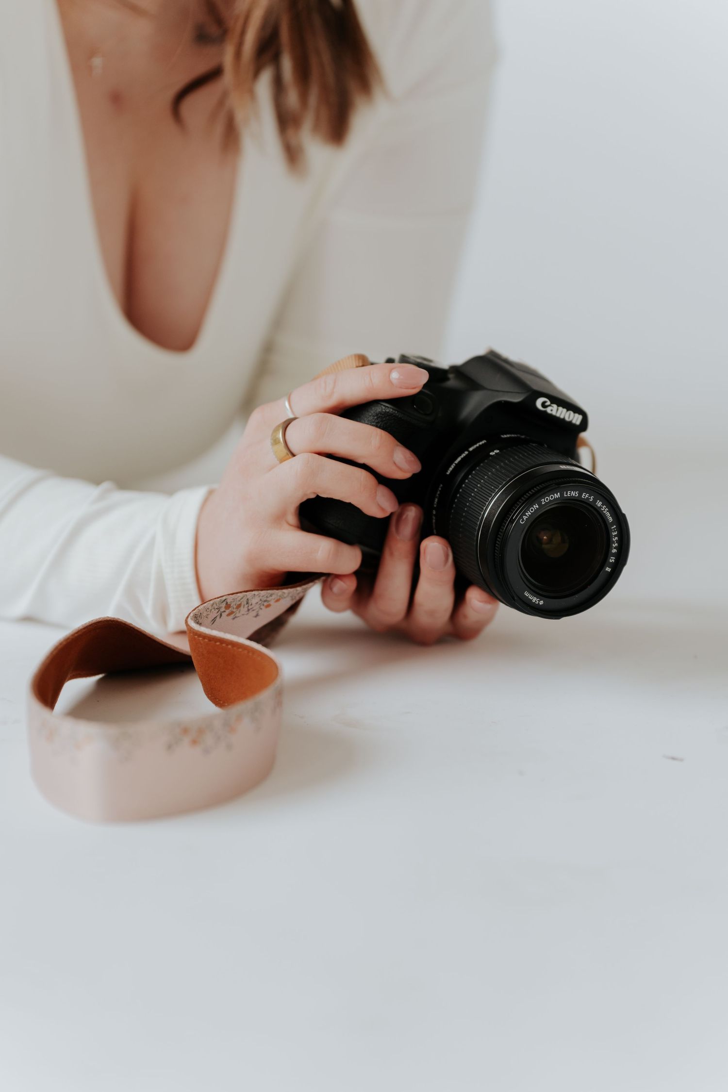 Woman in white top holding a Canon DSLR camera with a leather strap on a white surface.