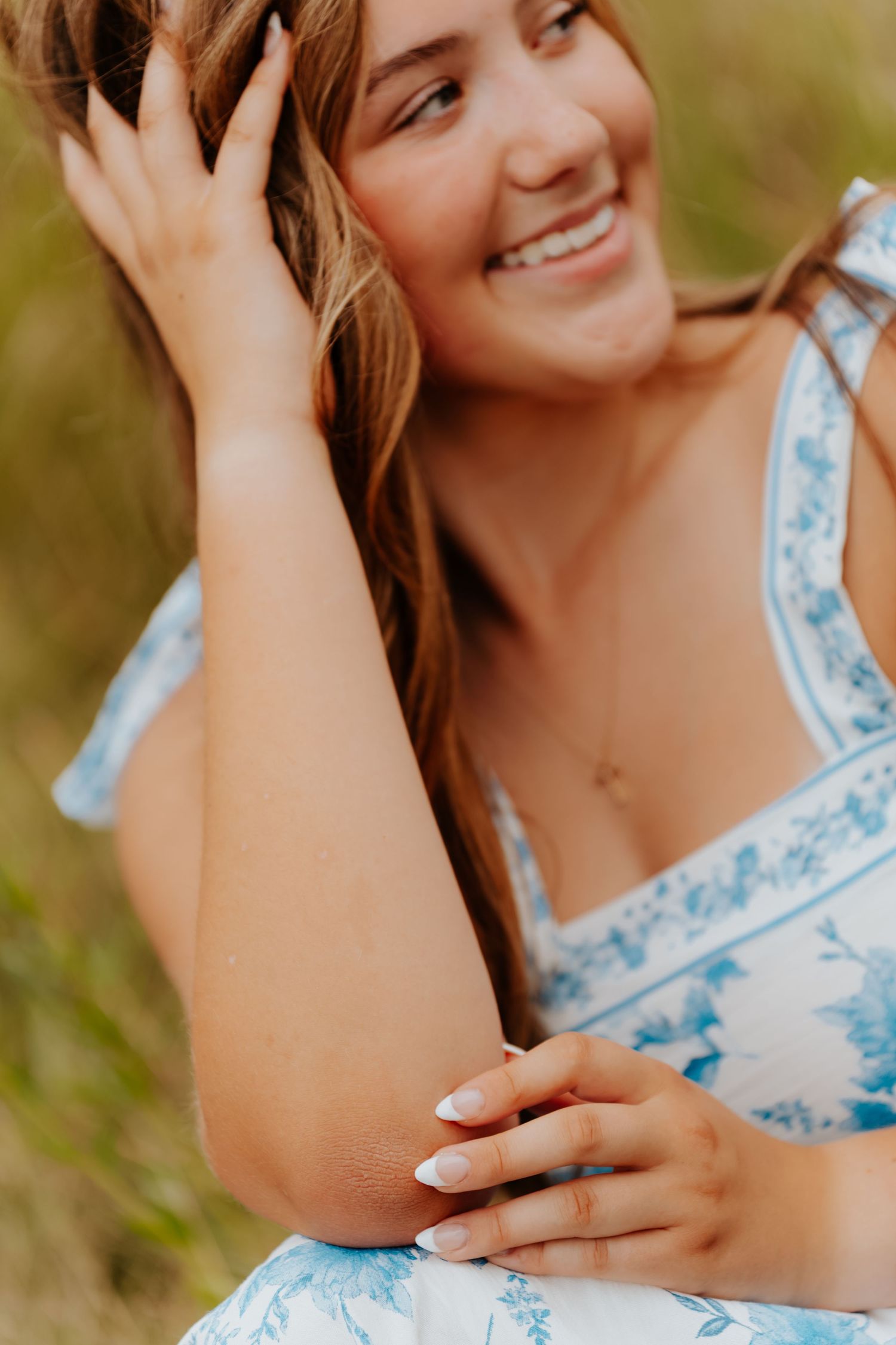 Smiling young woman in a blue floral dress sitting outdoors, hand in hair, with white manicured nails.
