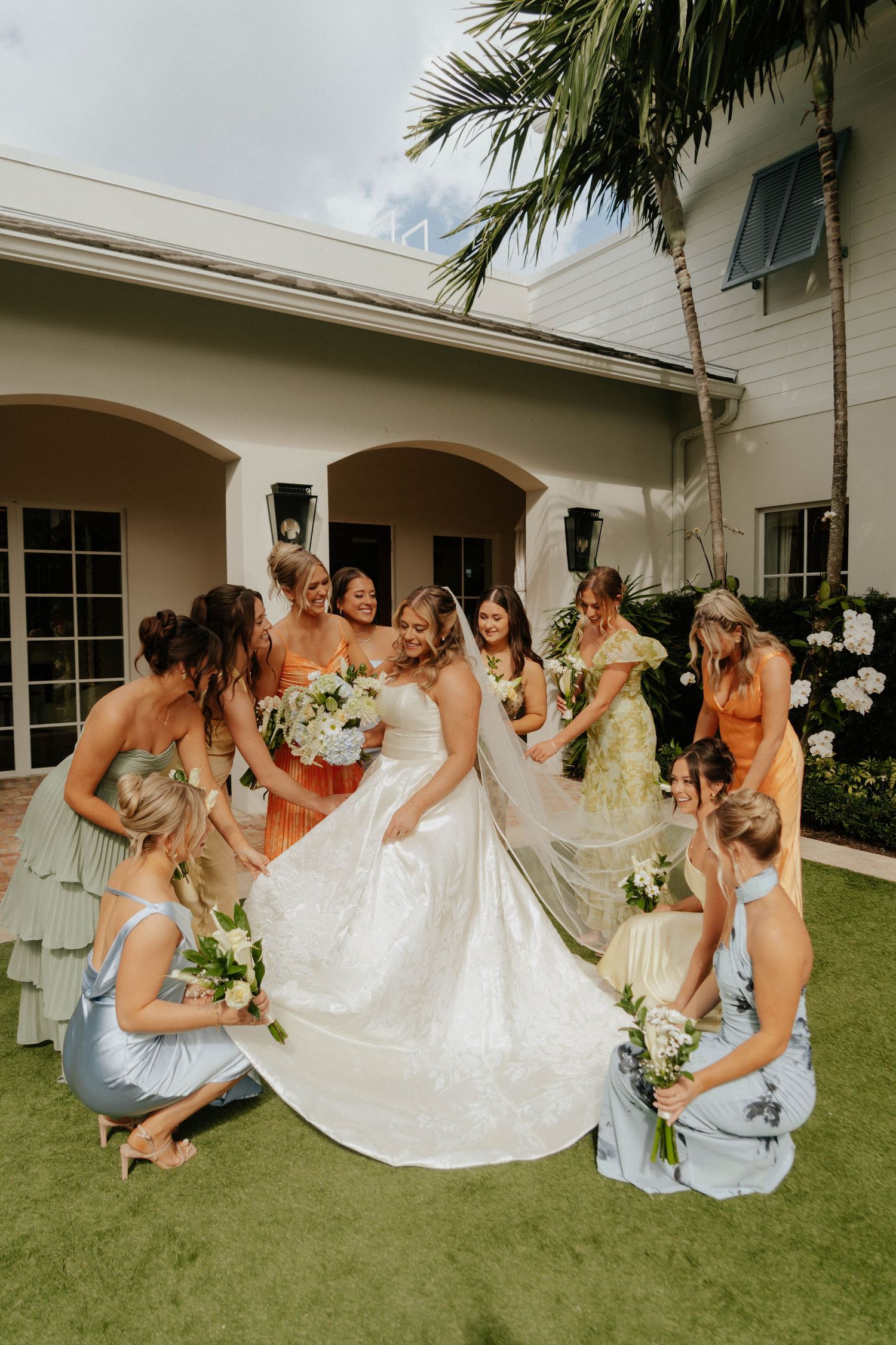 Bride surrounded by bridesmaids in colorful dresses holding white bouquets outdoors at a tropical wedding venue.