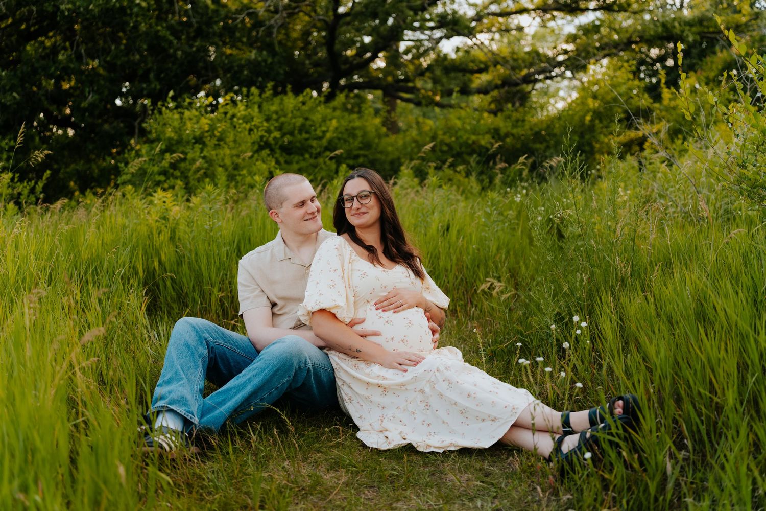Pregnant couple sitting in lush green field, woman in white dress cradling baby bump, man in jeans smiling at golden hour.