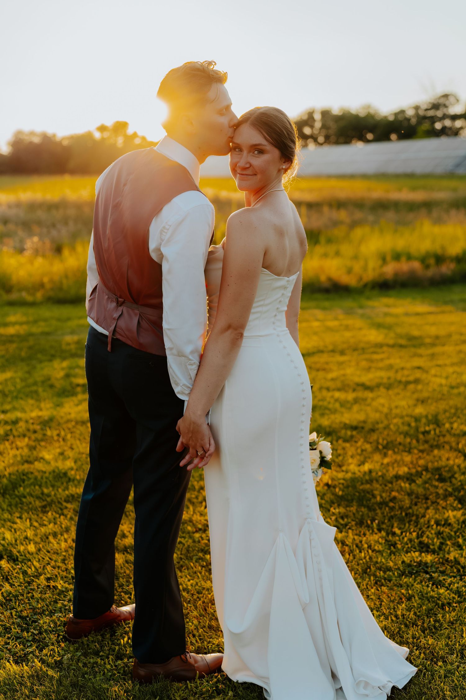 Bride and groom holding hands at golden hour sunset in a field, bride in white gown looks back smiling.