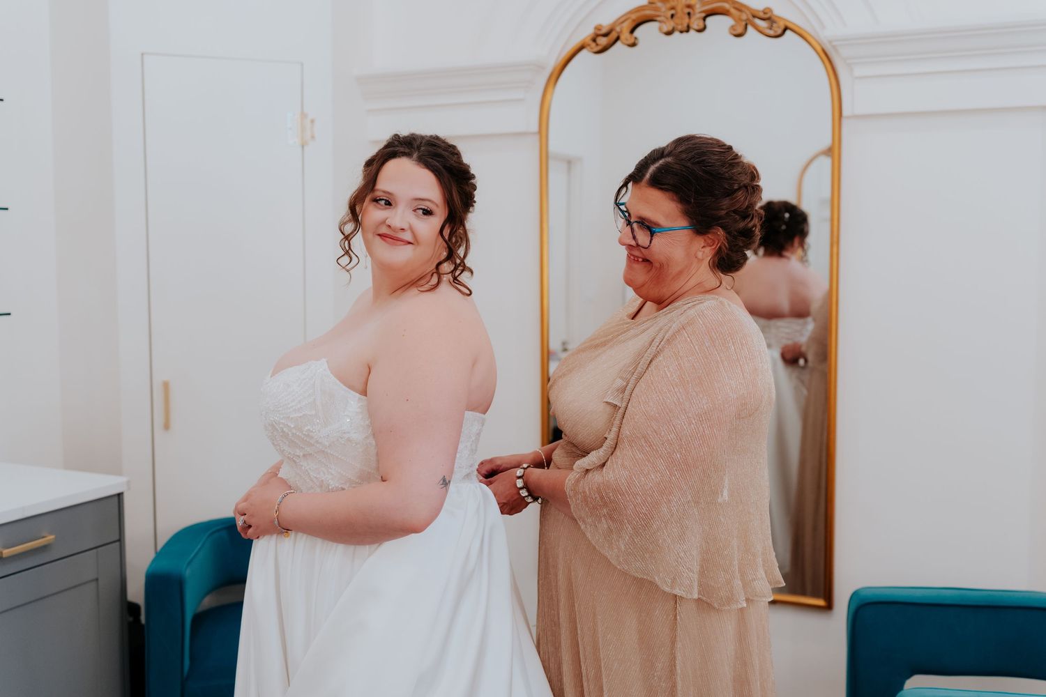 A bride in a white gown gets help fastening her dress from a woman in a gold top, standing before a gold mirror.