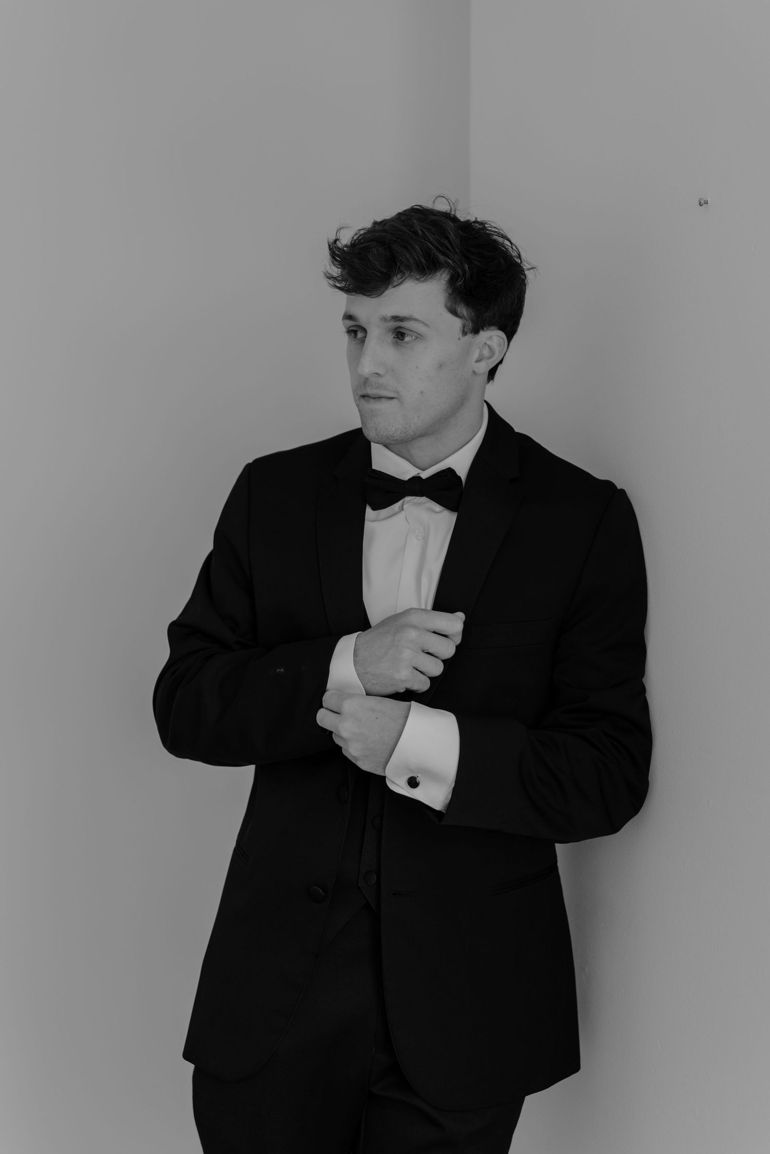 Black and white photo of a young man in a tuxedo with bow tie adjusting his cufflinks against a light wall.