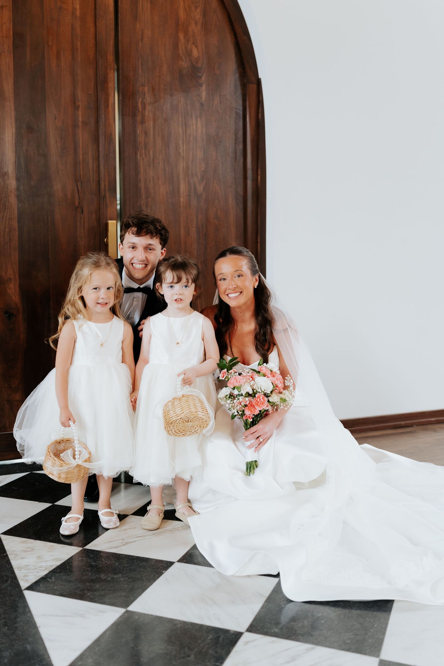 Bride and groom pose with two flower girls in white dresses on a checkered floor near wooden doors.