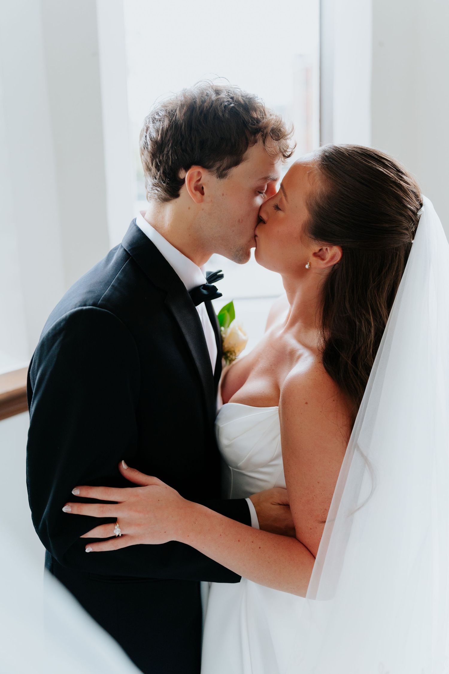 A bride and groom share a romantic kiss in a bright indoor setting, with the bride wearing a white veil.