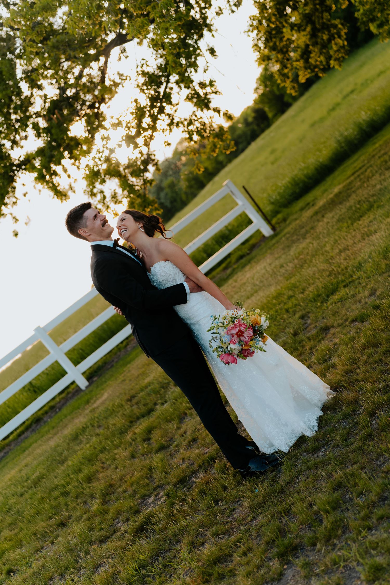Bride and groom embracing in a sunny green field by a white fence, bride holding a colorful bouquet in wedding attire.