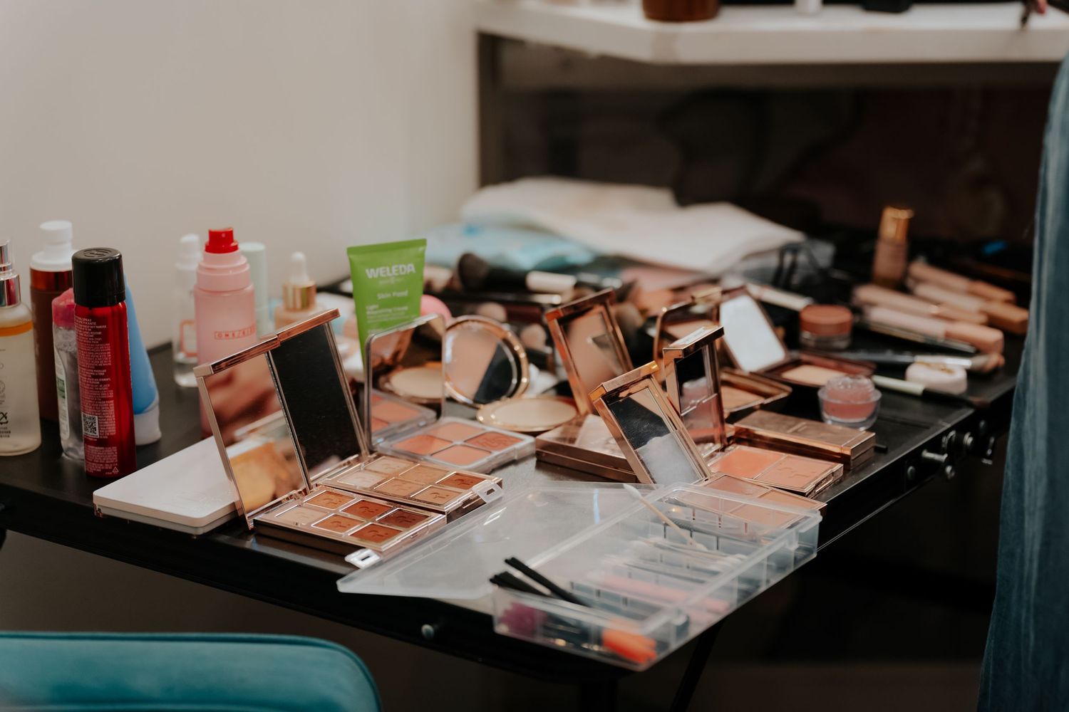 Cluttered makeup vanity table with rose gold eyeshadow palettes, mirrors, brushes, and beauty products.