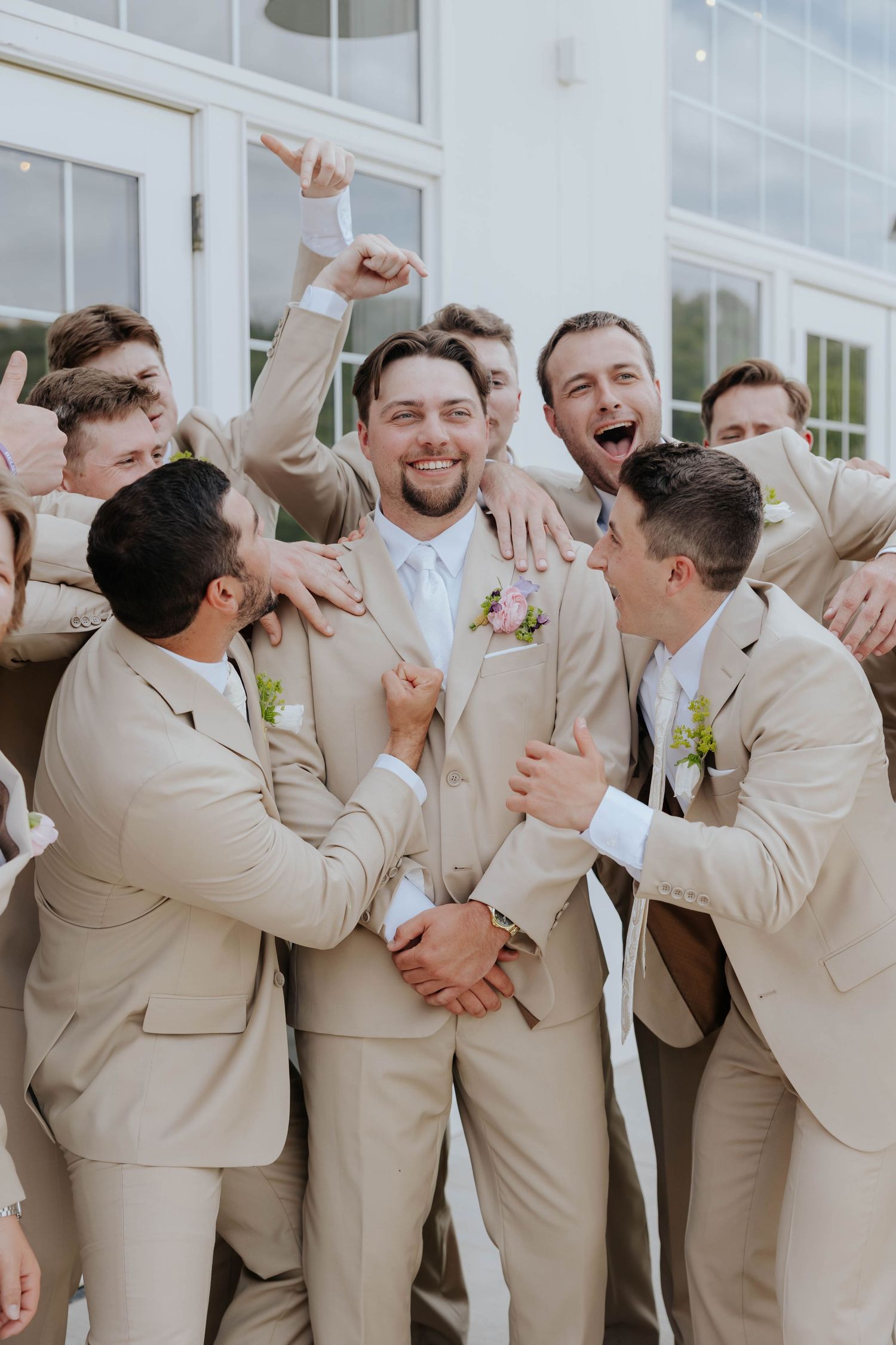 Groom in tan suit with boutonniere celebrates joyfully surrounded by cheering groomsmen in matching beige suits outdoors.