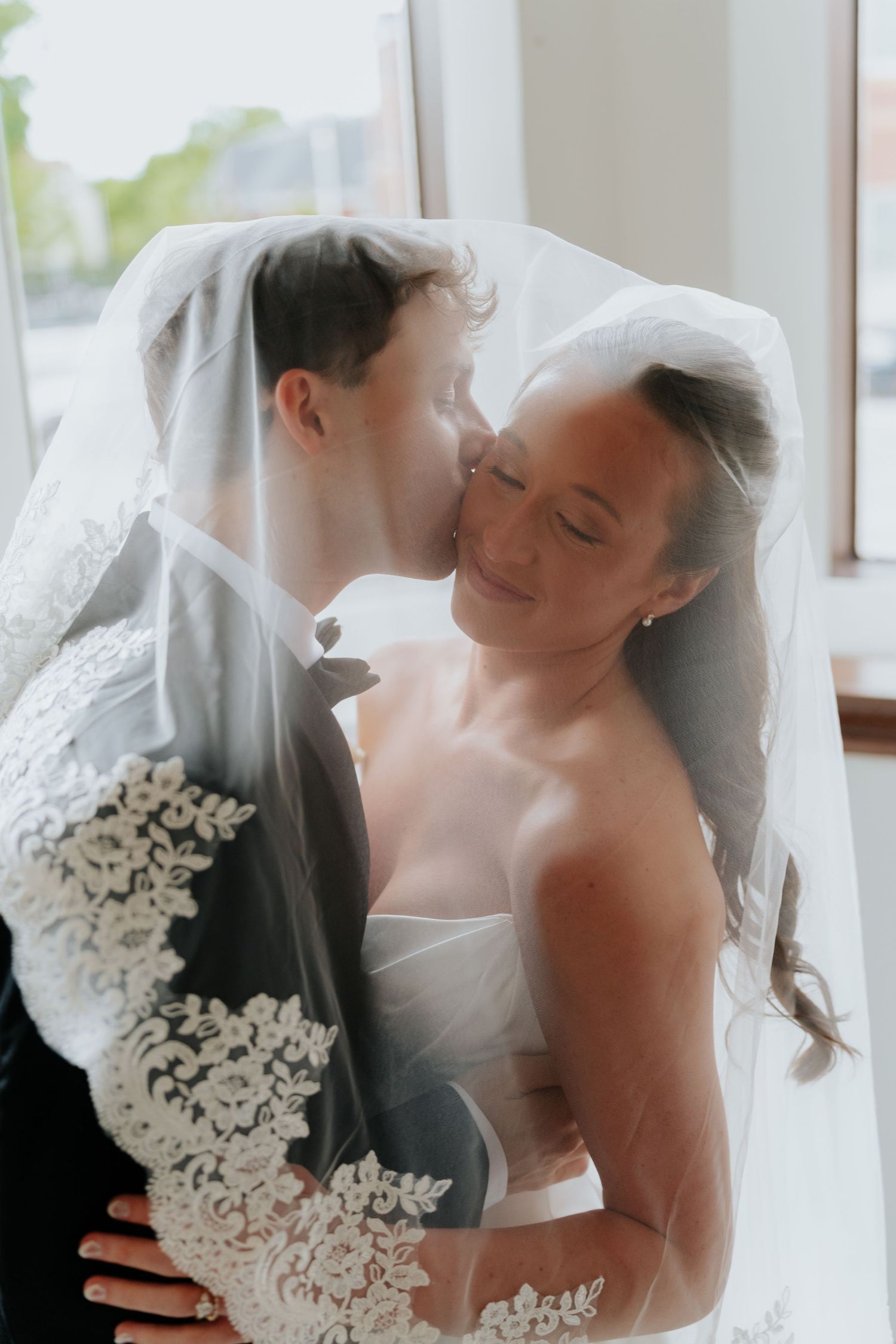 Bride smiles as groom kisses her cheek under a lace-trimmed veil in a romantic wedding portrait.