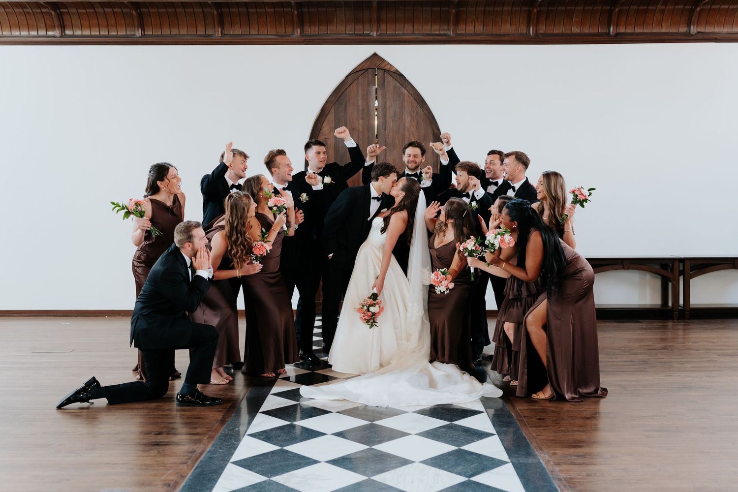 Wedding party celebrating on checkered dance floor with bridesmaids in brown dresses and groomsmen in black tuxedos.