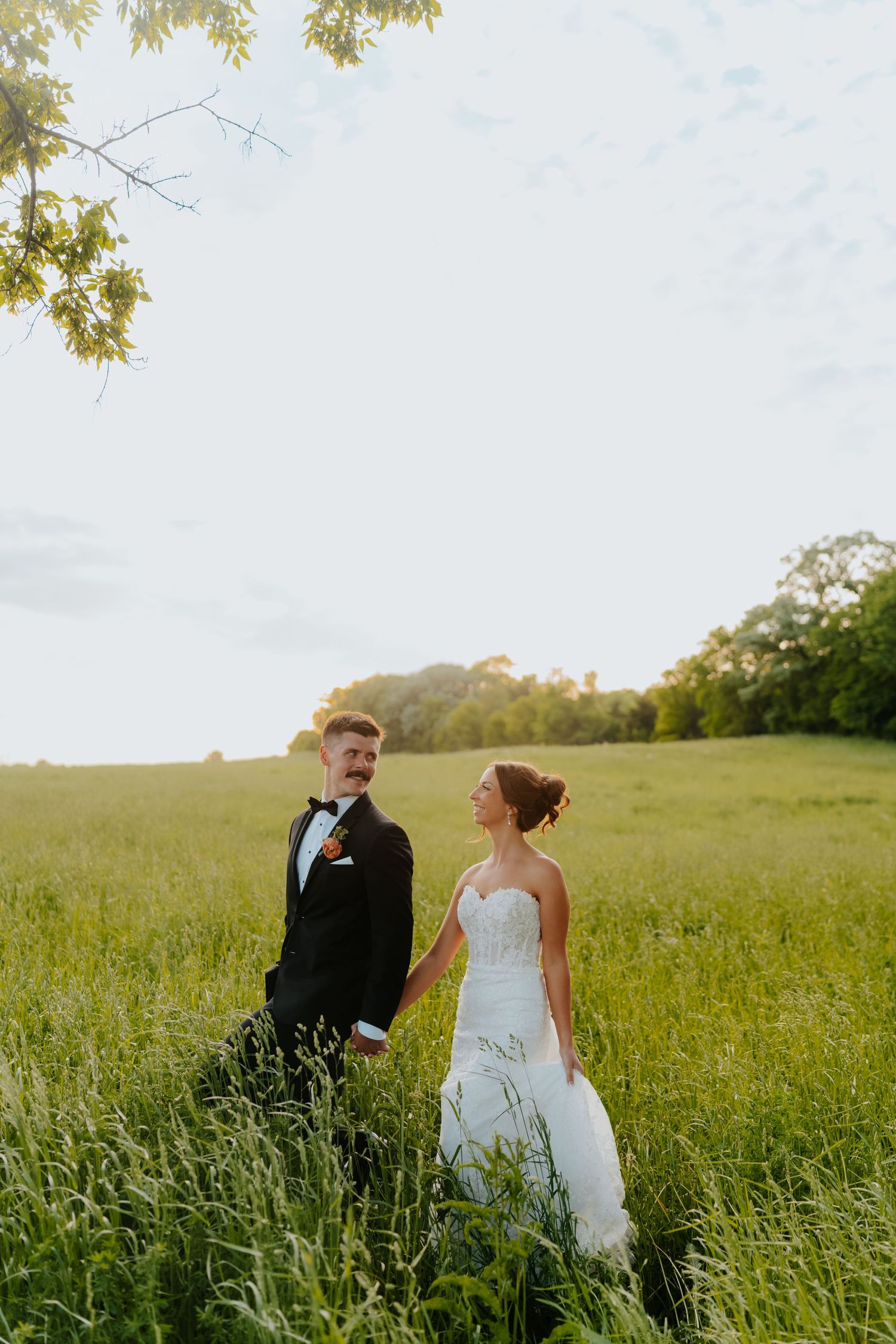 Bride and groom holding hands in a lush green field at golden hour, bride in white gown, groom in black tuxedo.